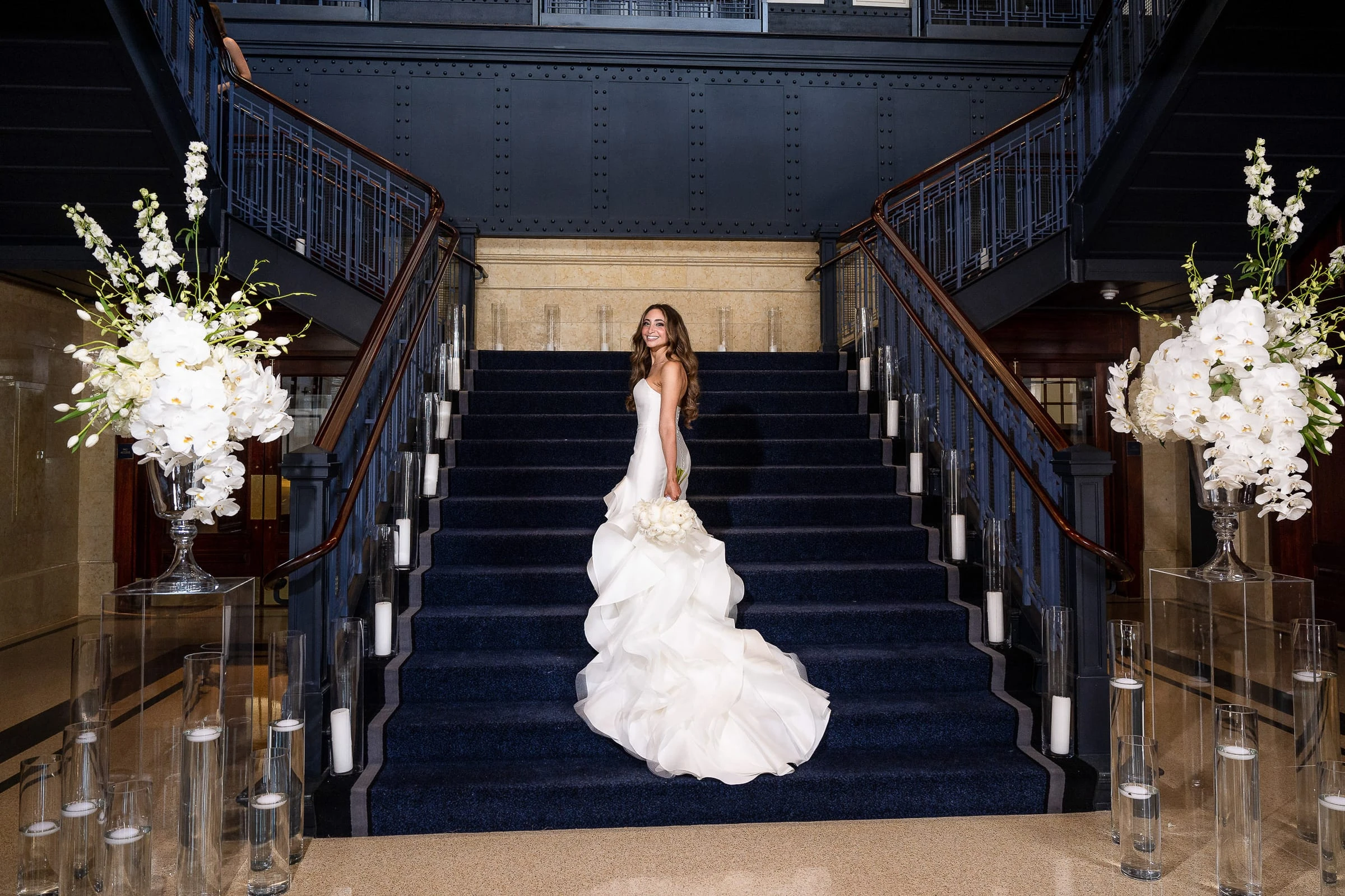 The bride alone on the blue staircase between tall white orchids — Casa Cipriani, New York