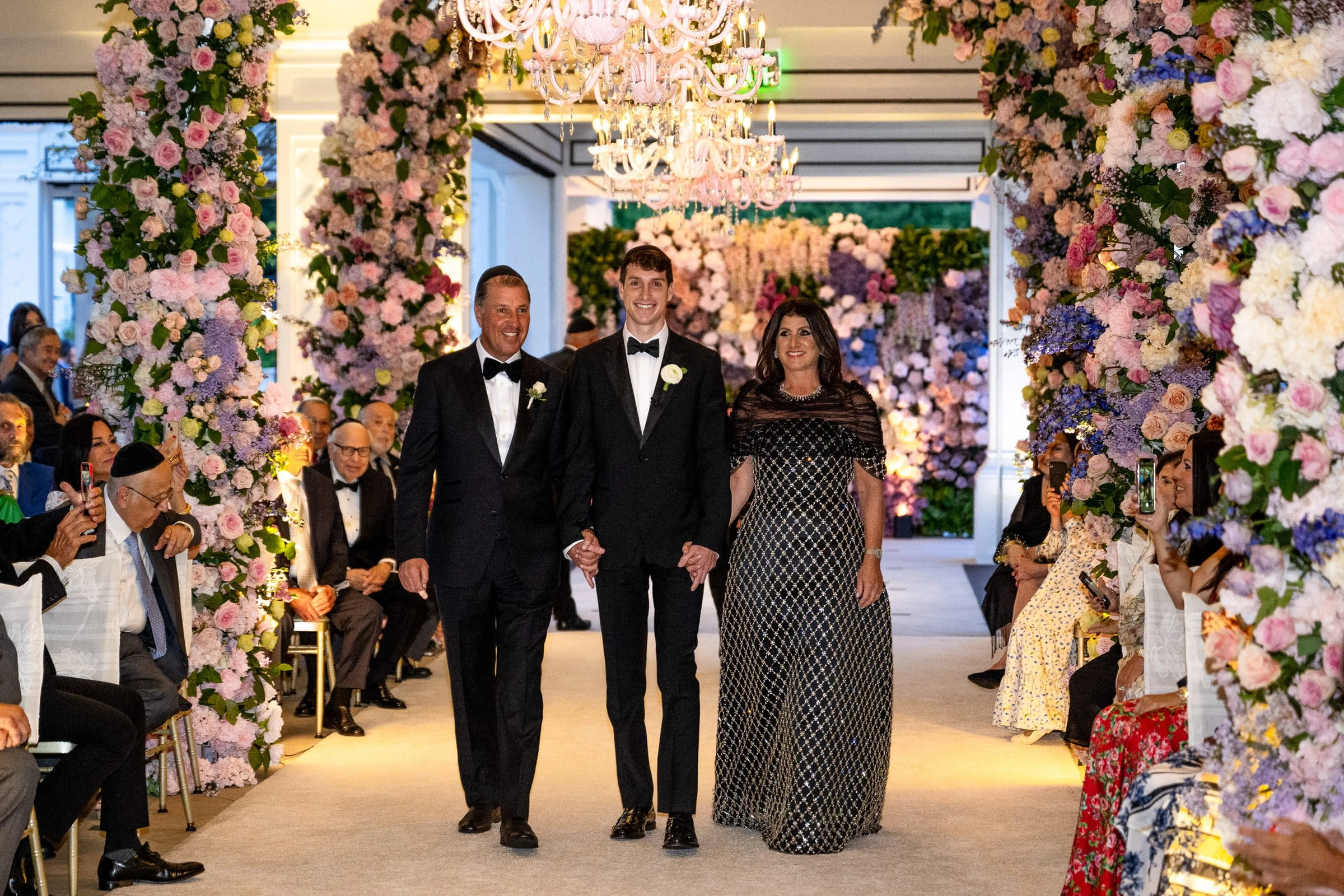 The groom walking the aisle between his parents beneath the flower arch — Shadowbrook at Shrewsbury, Shrewsbury