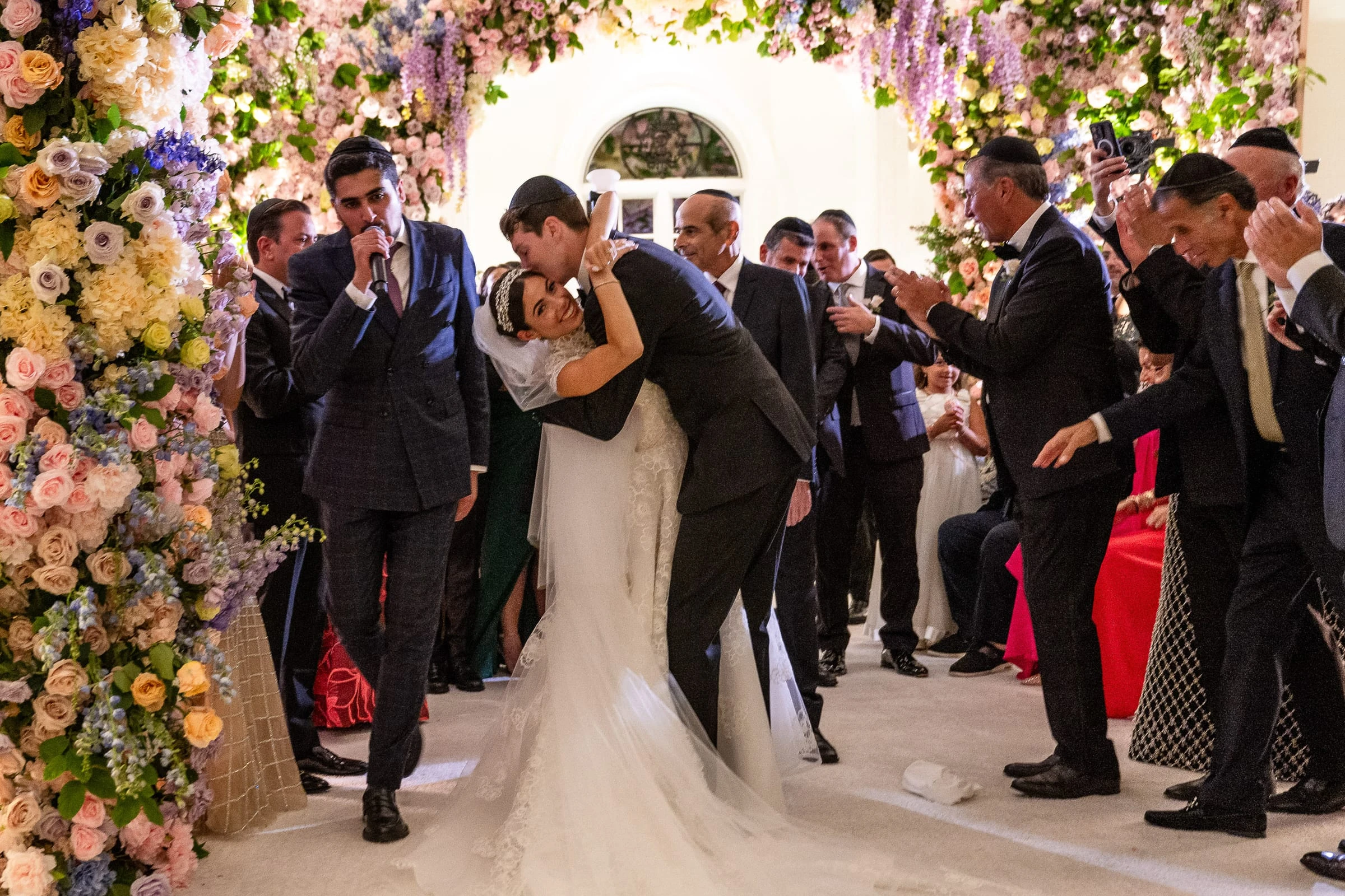 The groom dipping the bride for a kiss beneath the flowered chuppah as guests cheer — Shadowbrook at Shrewsbury, Shrewsbury
