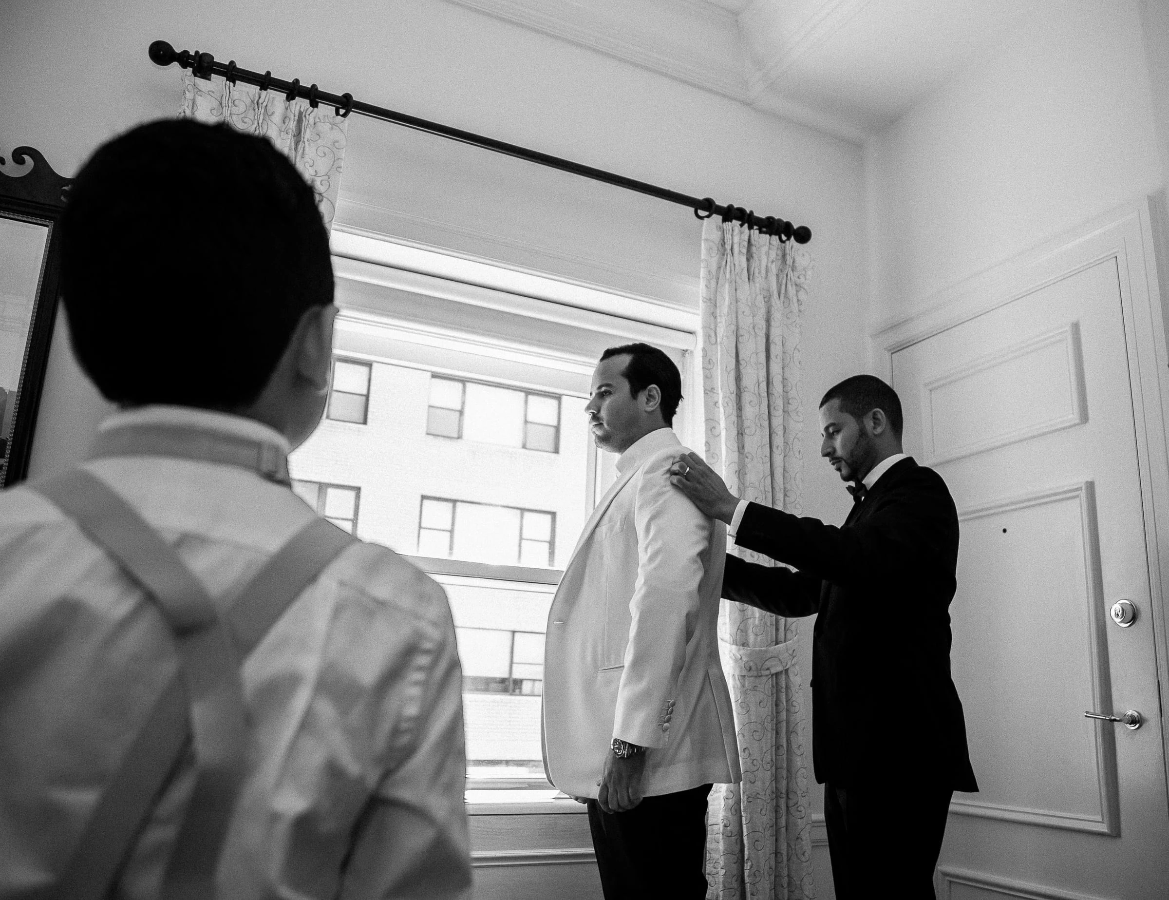A groom having his white jacket straightened at the window, in black and white — Metropolitan Club, New York