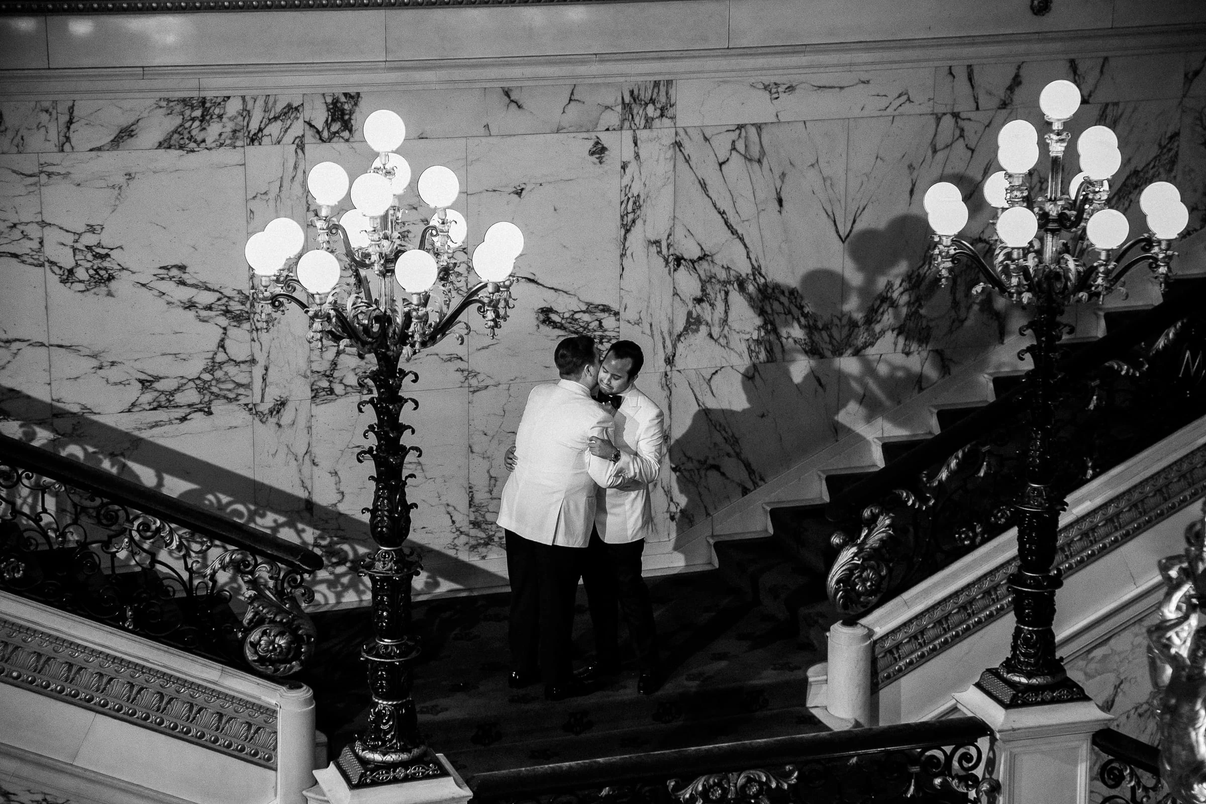 The two grooms embracing on the marble stairs between the candelabra, in black and white — Metropolitan Club, New York