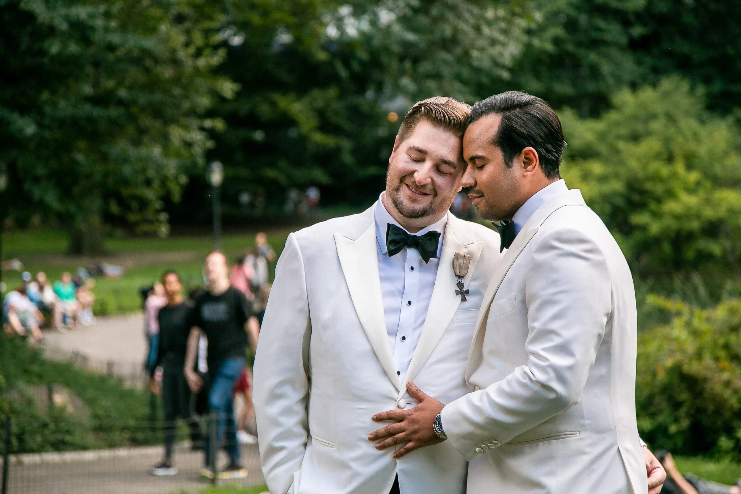 The grooms together in the park with the crowd passing behind — Metropolitan Club, New York
