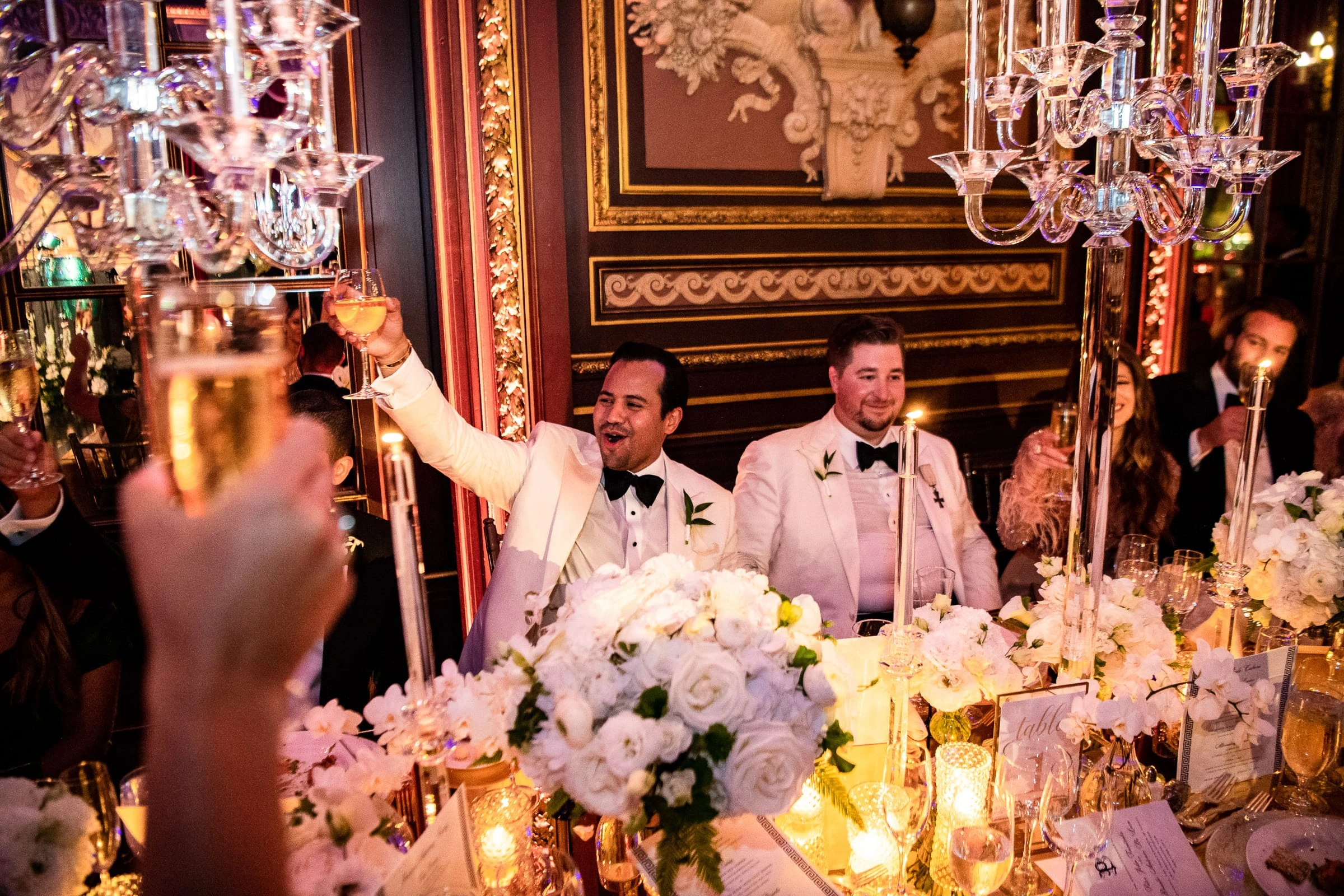 The two grooms raising their glasses for a toast at the candlelit head table — Metropolitan Club, New York