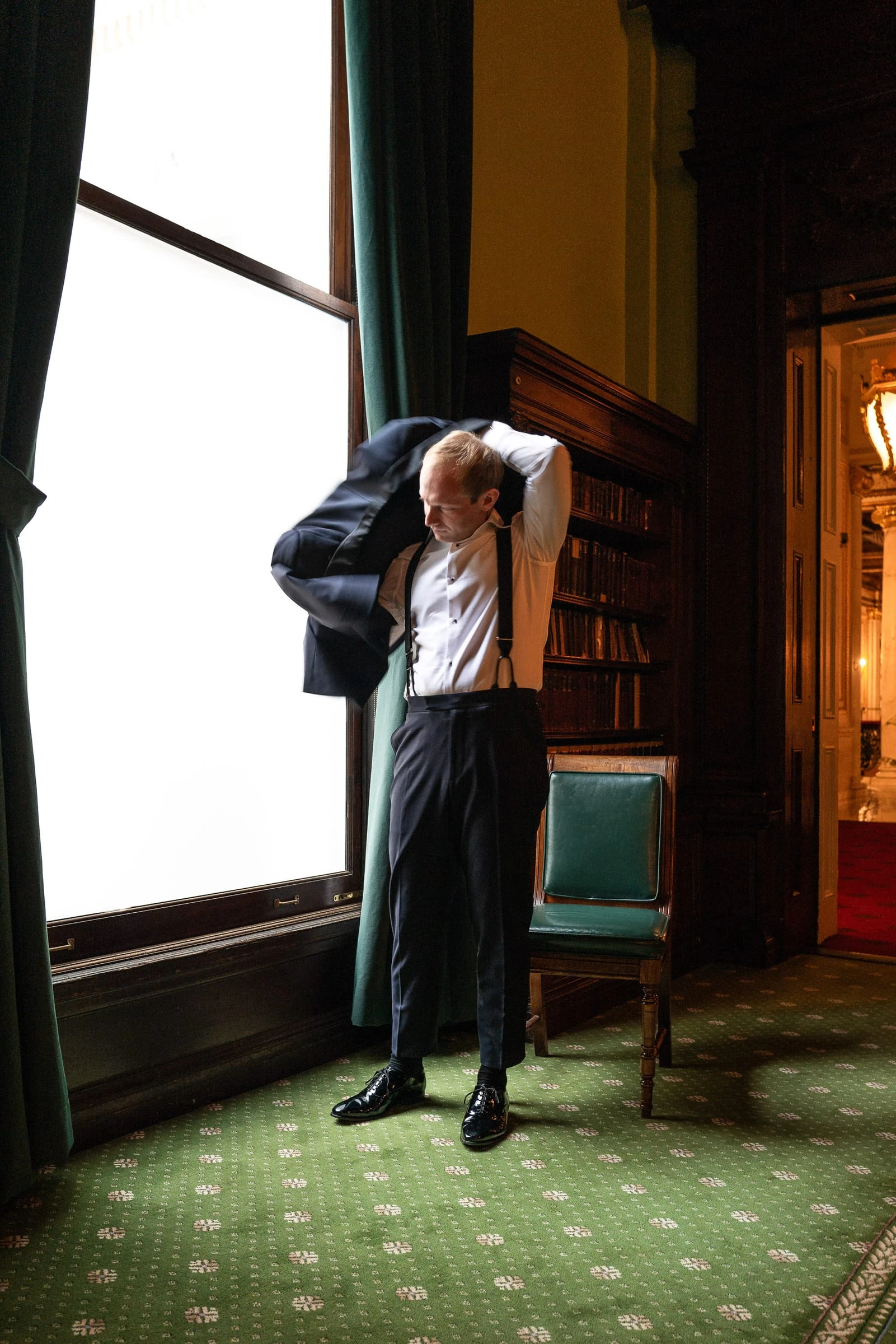 The groom pulling on his jacket at the library window — Metropolitan Club, New York