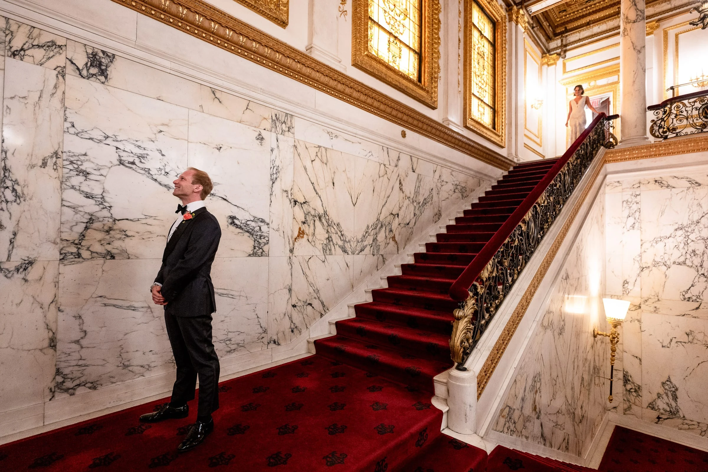 The groom looking up the marble staircase as the bride appears at the top — Metropolitan Club, New York