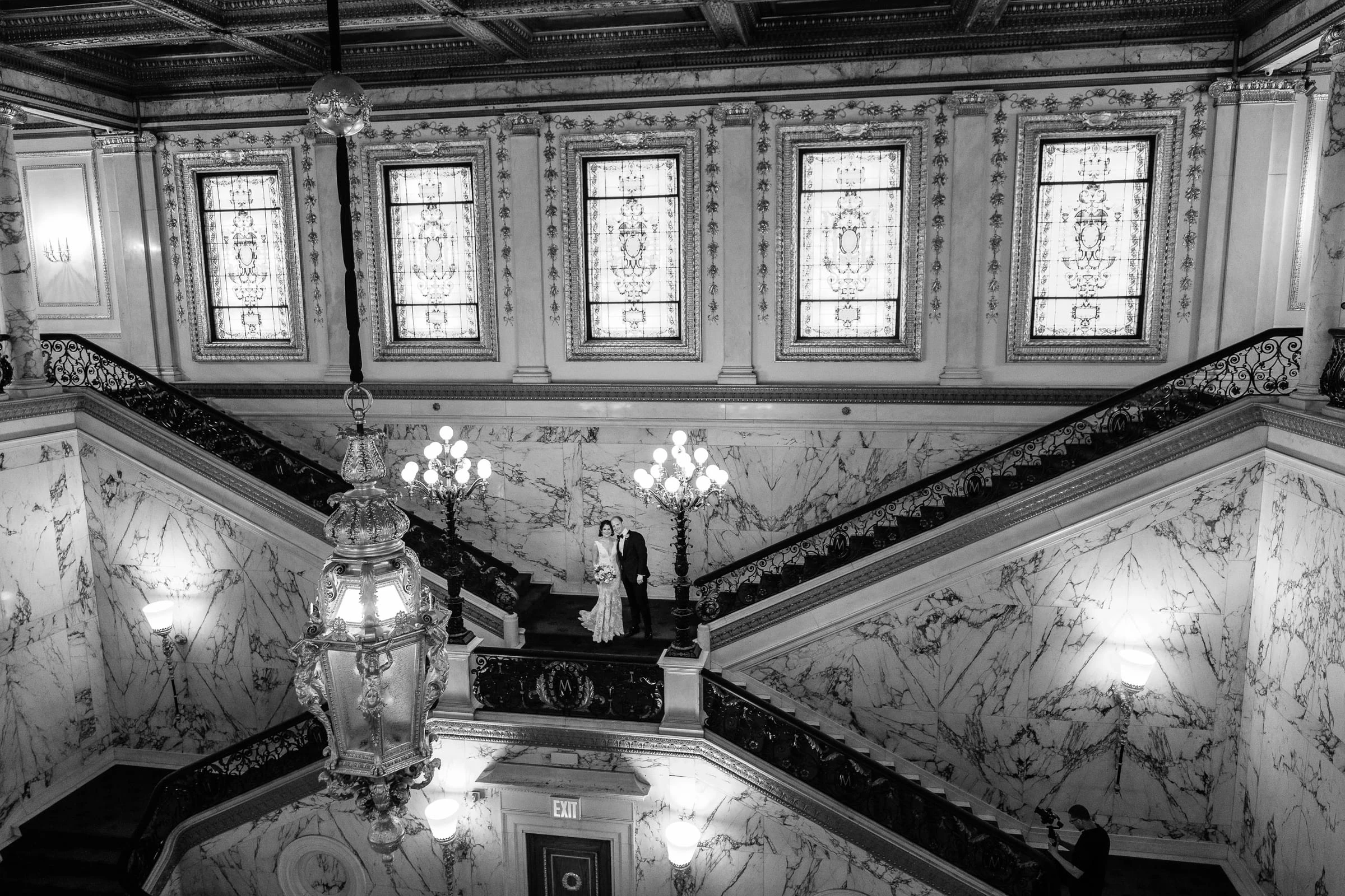 The couple small in the centre of the stair hall beneath its lanterns, in black and white — Metropolitan Club, New York
