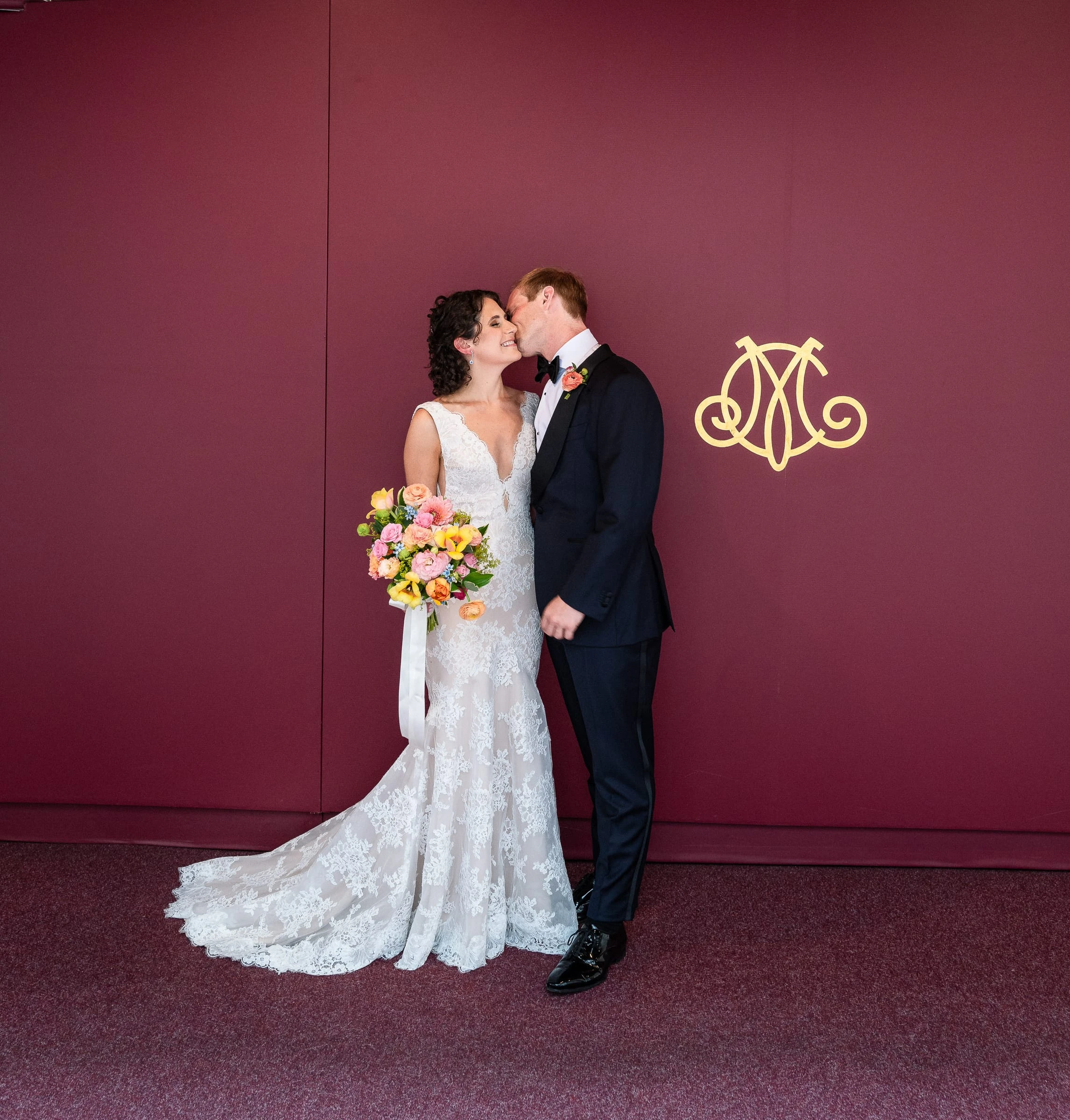 The couple kissing against a deep red wall marked with the club monogram — Metropolitan Club, New York