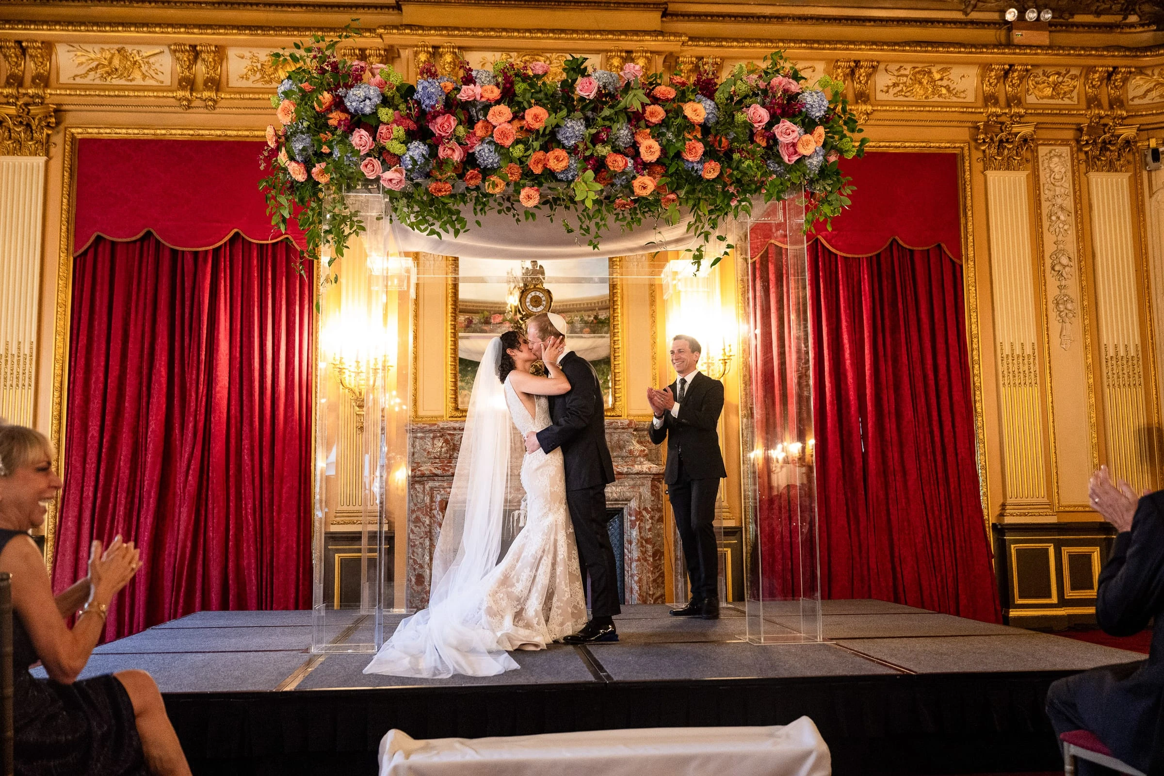 The couple kissing beneath the flowered chuppah as guests applaud — Metropolitan Club, New York