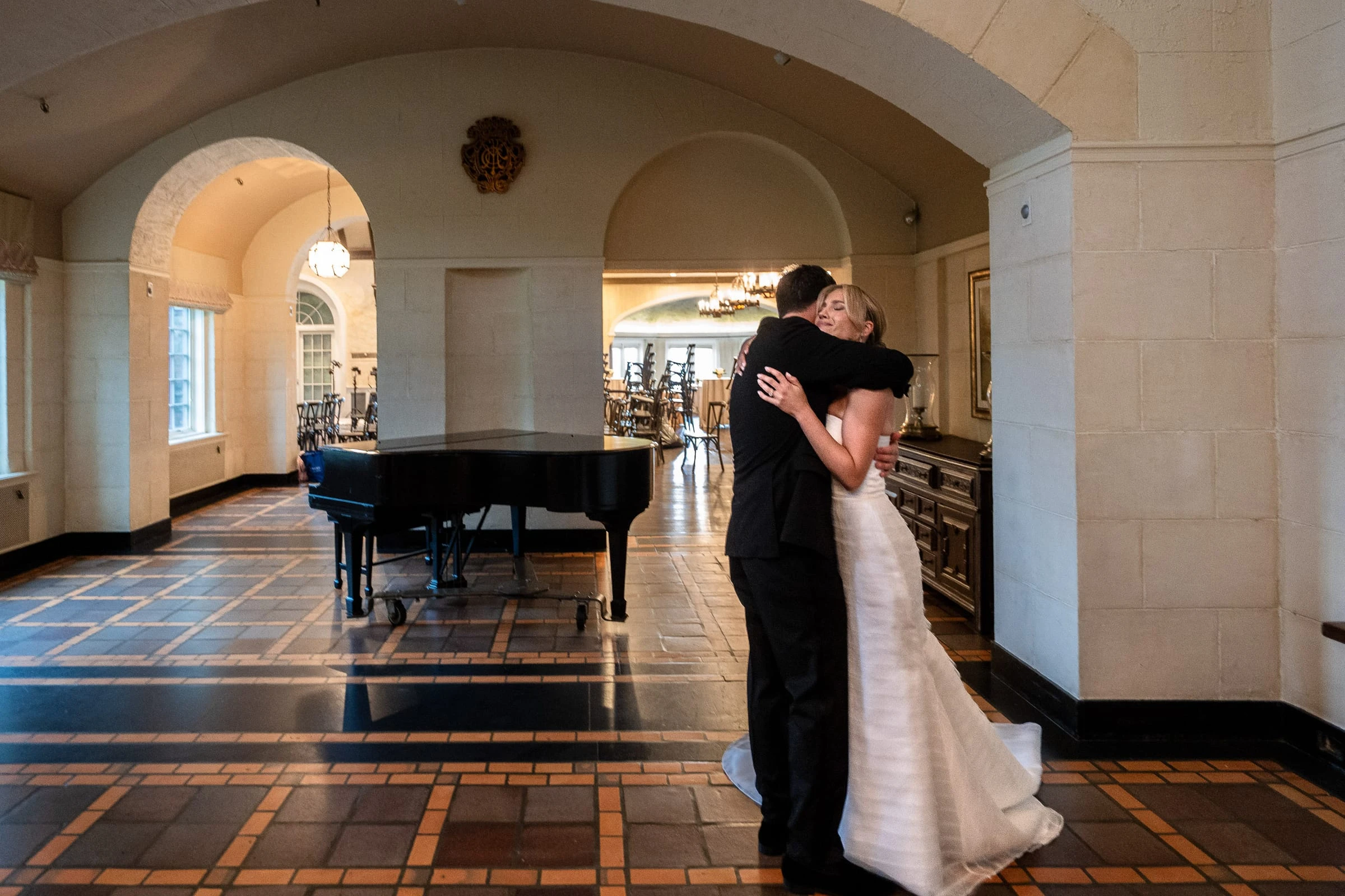 The couple embracing in the tiled hall beside the grand piano — Mountain Ridge Country Club, West Caldwell