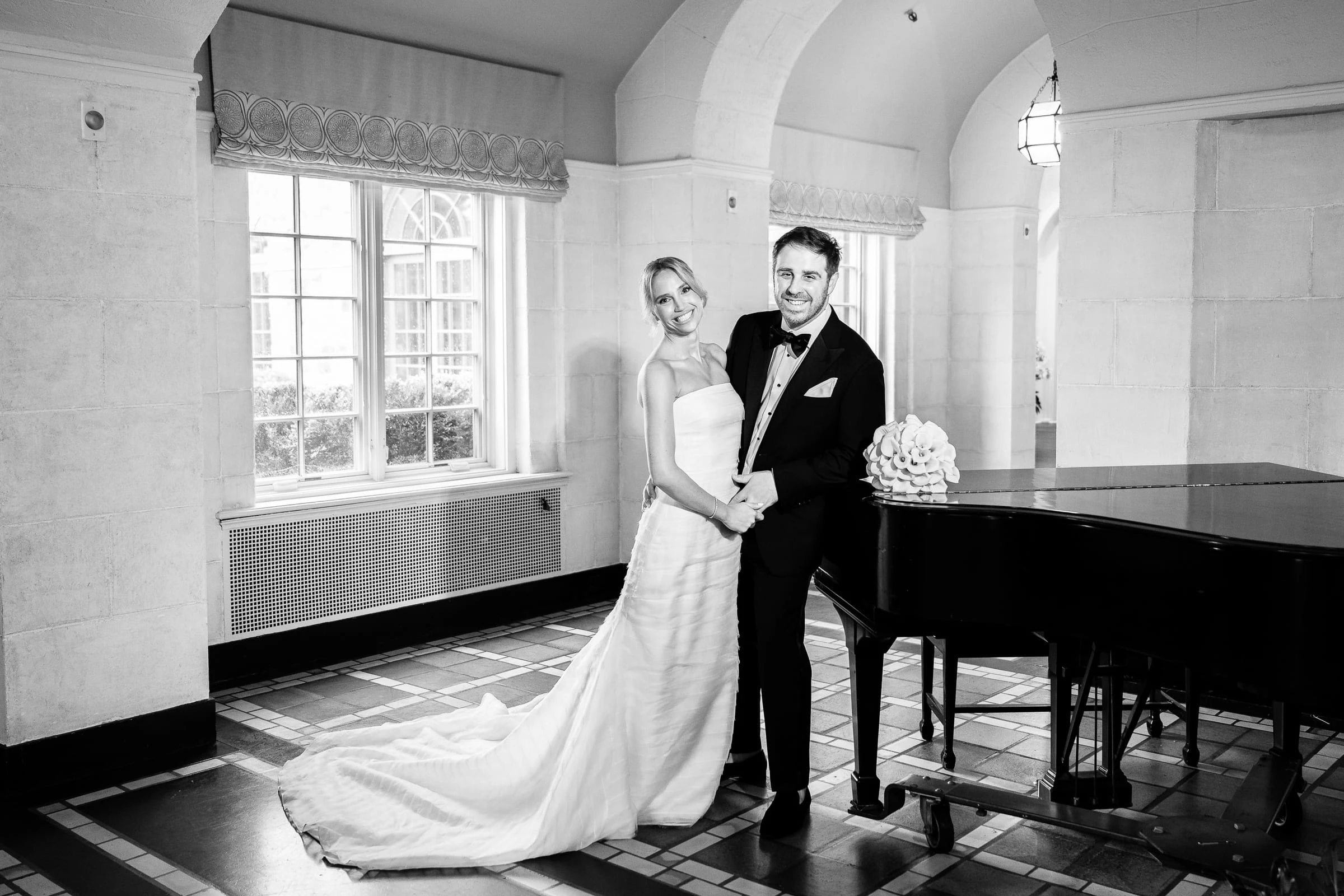 The couple beside the grand piano in the stone-arched room, in black and white — Mountain Ridge Country Club, West Caldwell