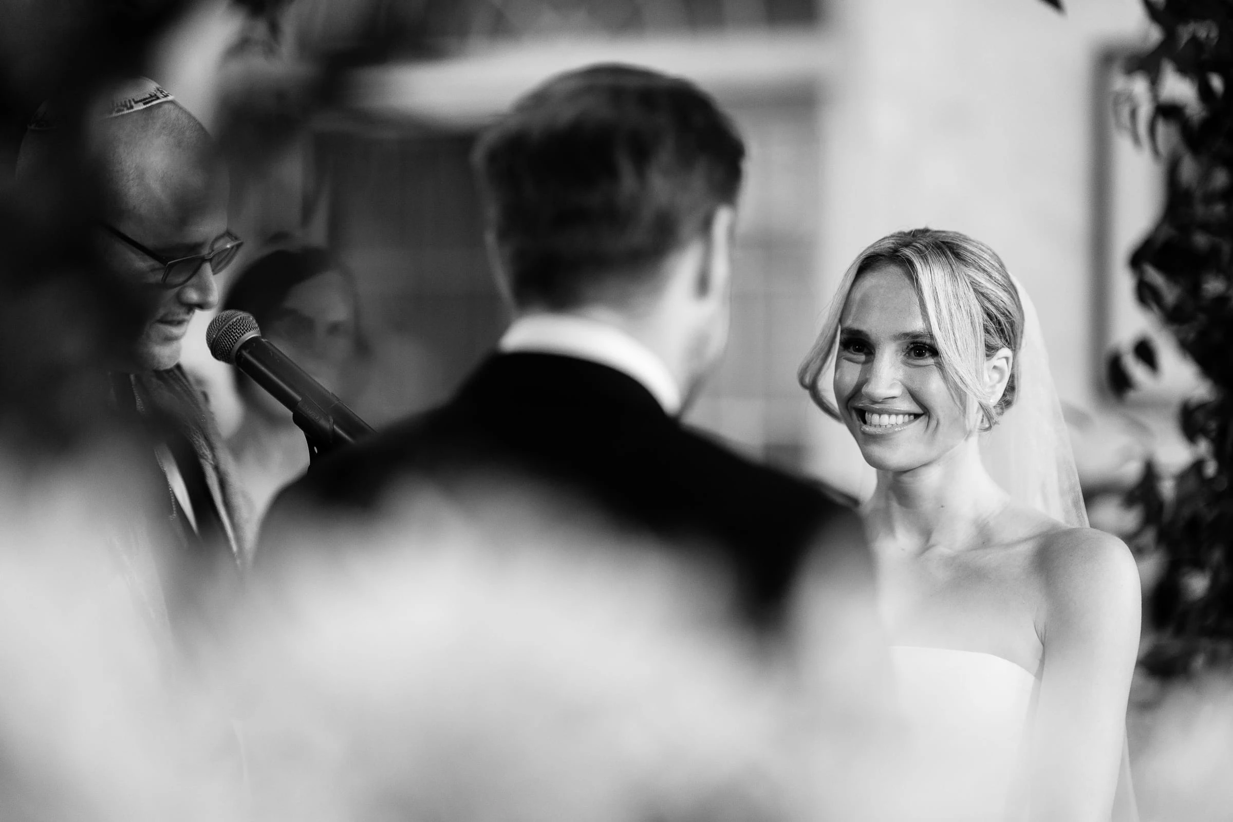 The bride smiling at the groom during the ceremony, framed through soft-focus flowers, in black and white — Mountain Ridge Country Club, West Caldwell