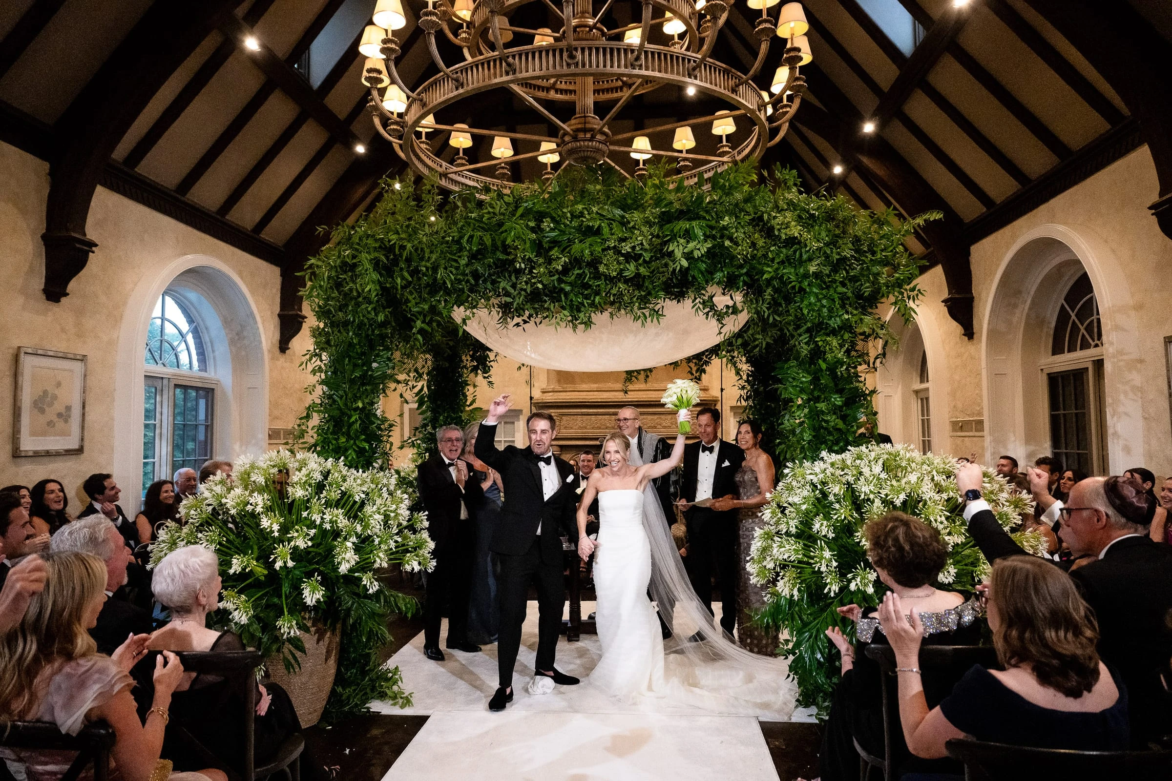 The couple walking back up the aisle beneath a canopy of greenery as guests applaud — Mountain Ridge Country Club, West Caldwell
