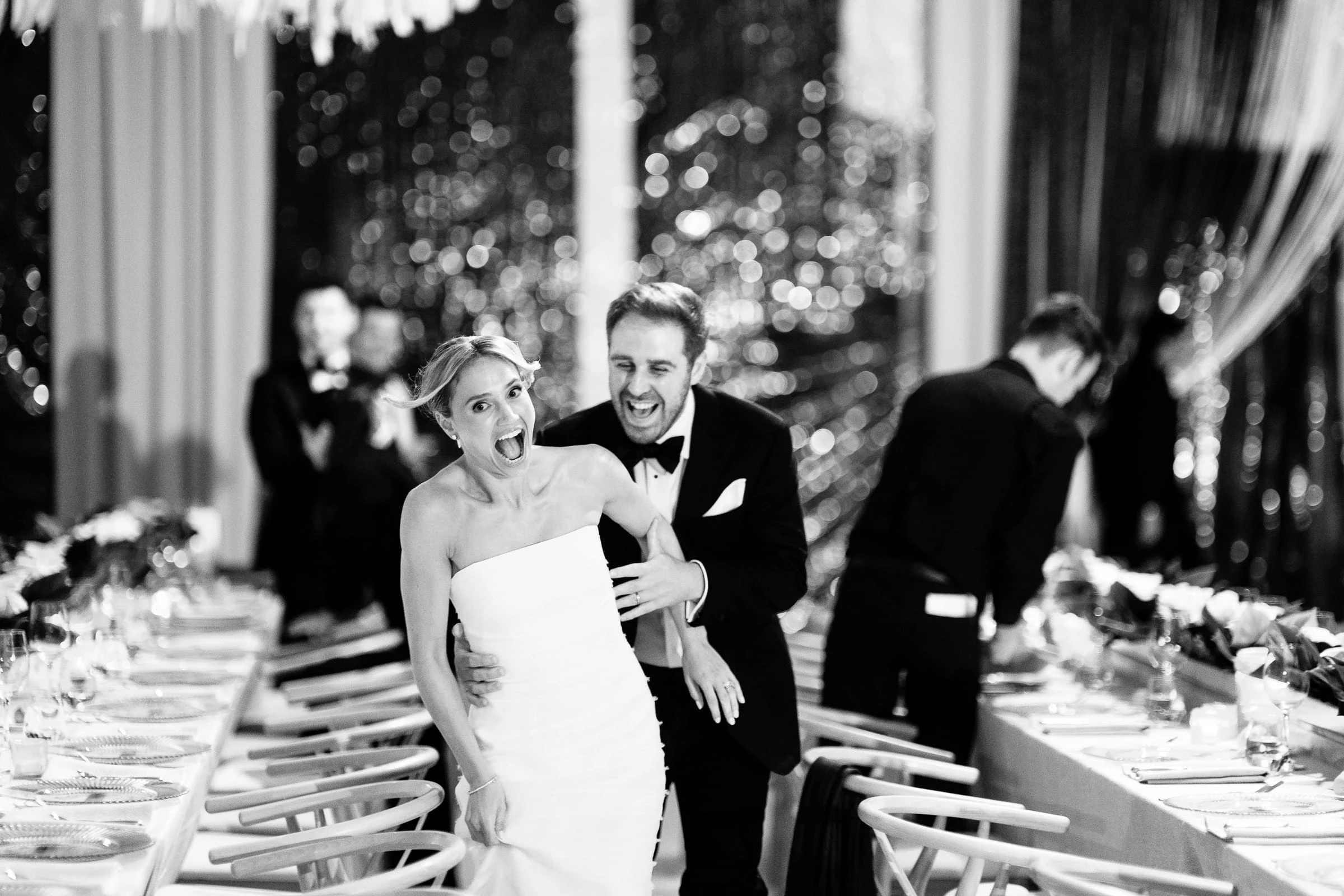 The bride and groom laughing as they make their entrance past the dinner tables, in black and white — Mountain Ridge Country Club, West Caldwell