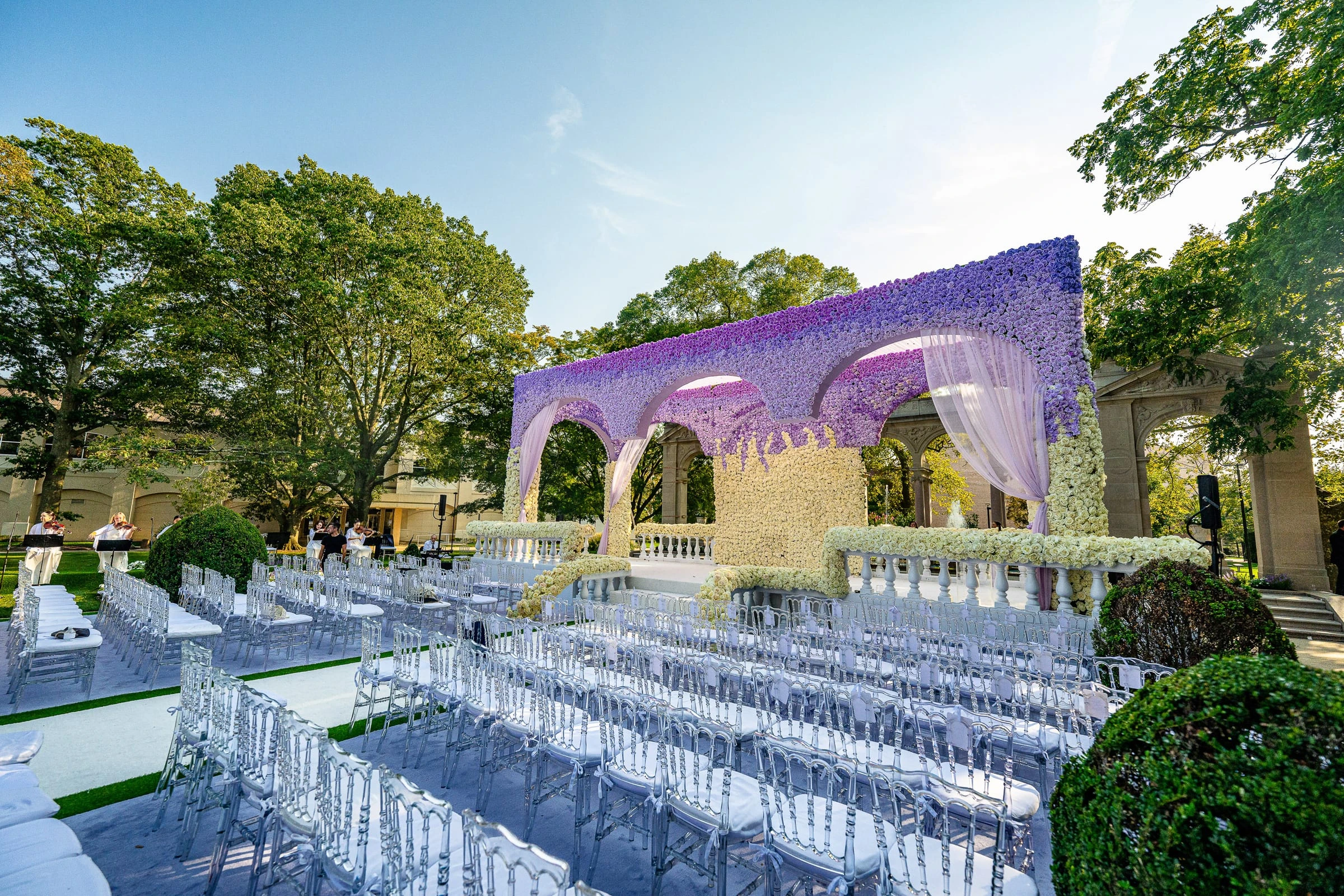 Rows of clear chairs on a lawn facing a lavender floral arcade set for an outdoor ceremony