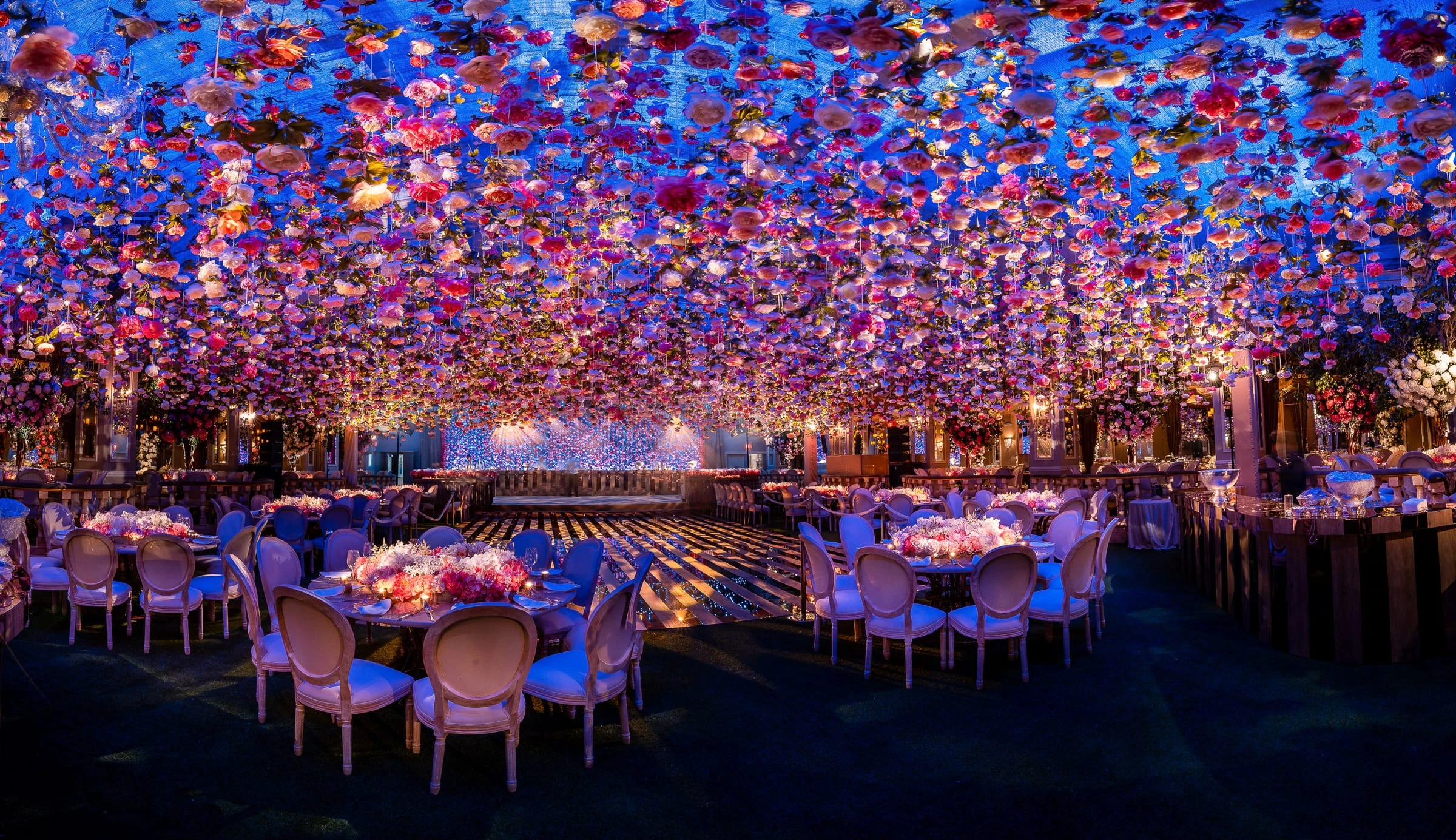 A ballroom of round tables beneath a vast ceiling of suspended flowers lit in blue and pink