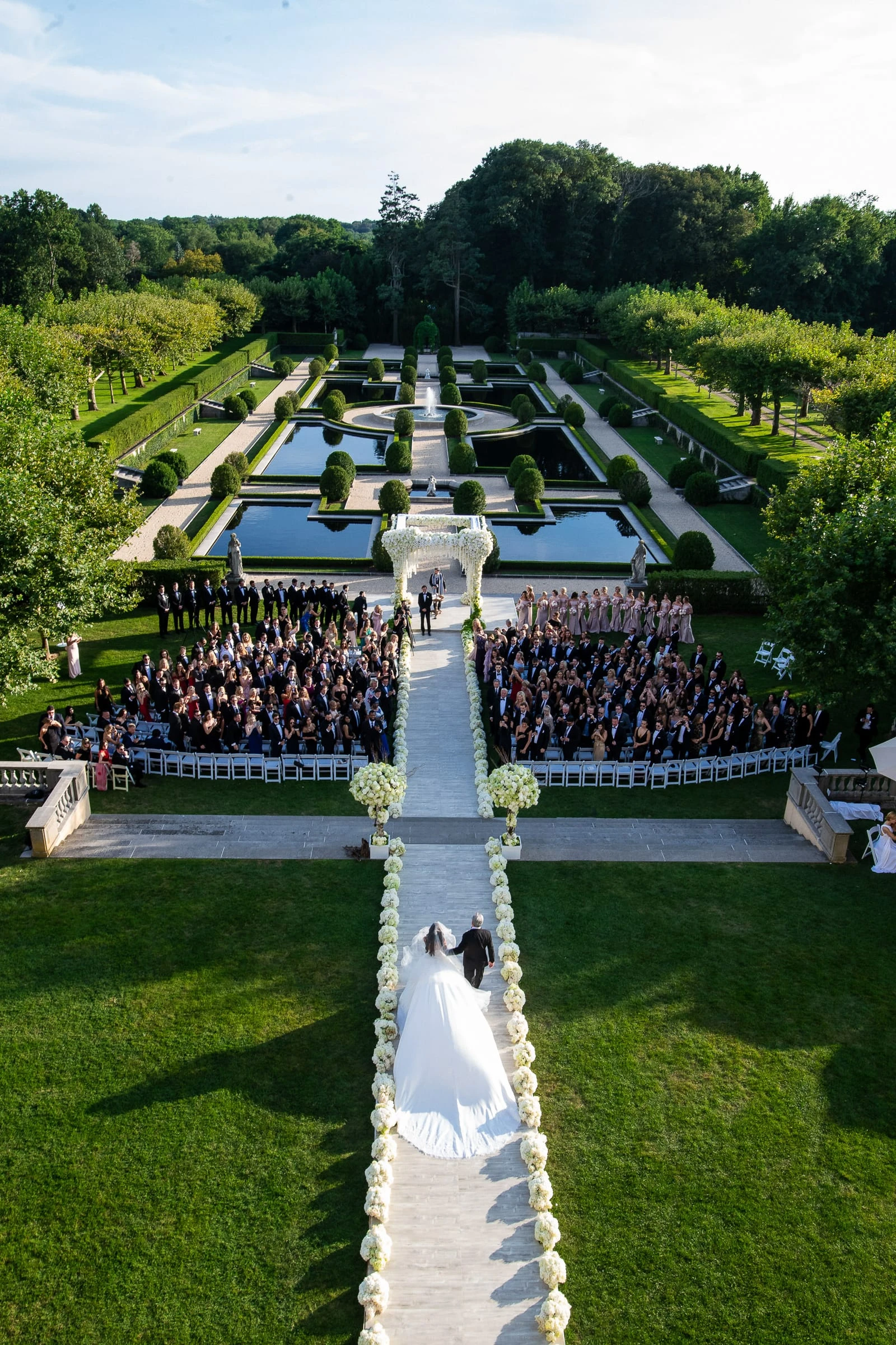 An outdoor ceremony from above, guests seated either side of a long aisle in formal gardens