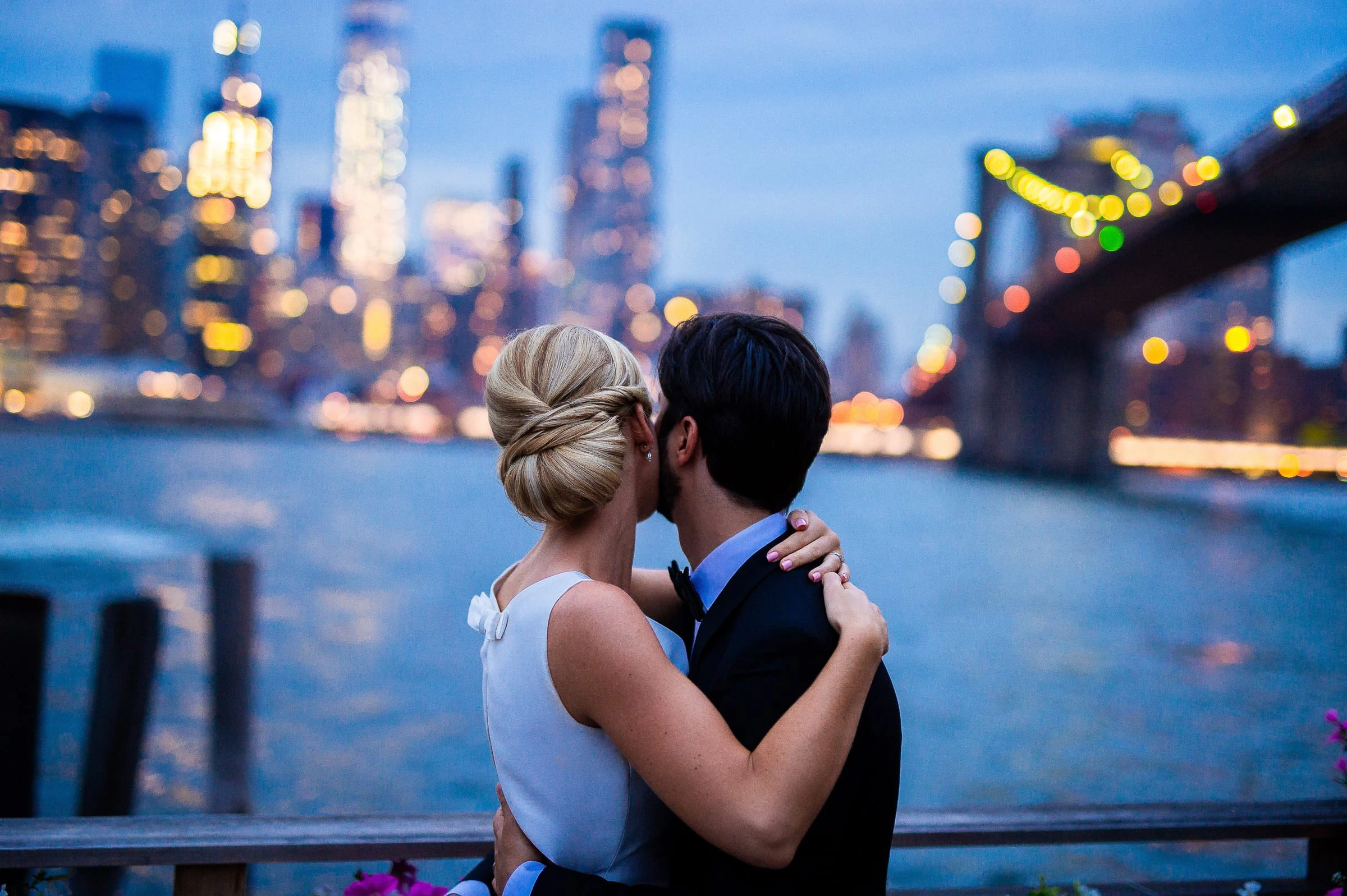 A couple kissing at a railing at dusk against the lit Manhattan skyline and the Brooklyn Bridge