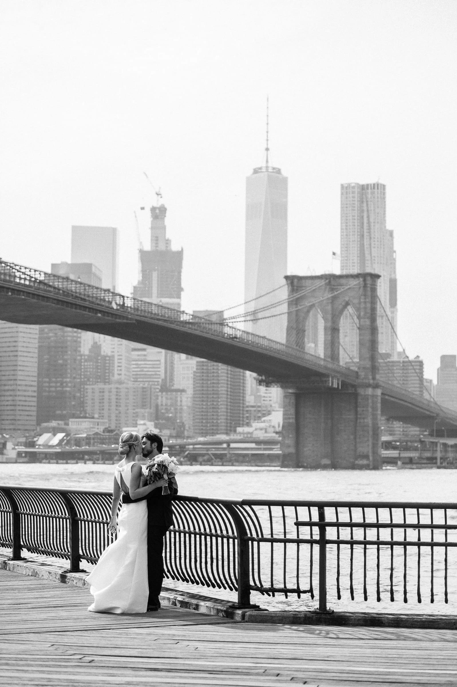 A couple embracing on the Brooklyn waterfront with the Brooklyn Bridge and the Manhattan skyline behind them, in black and white