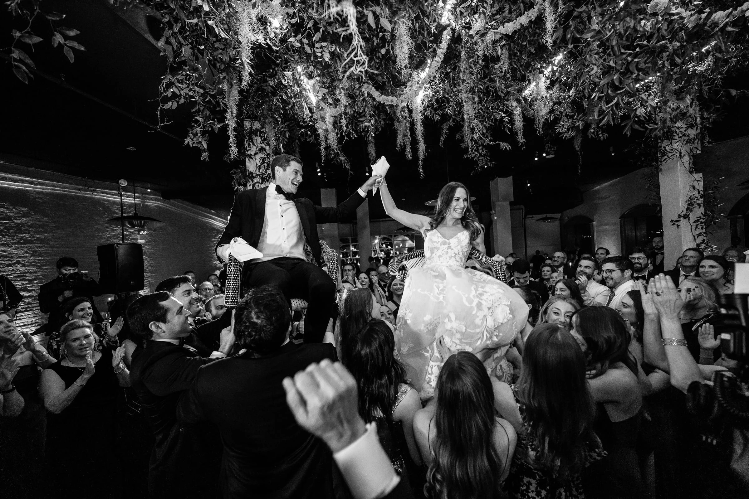The couple raised on chairs during the hora above a packed dance floor, in black and white