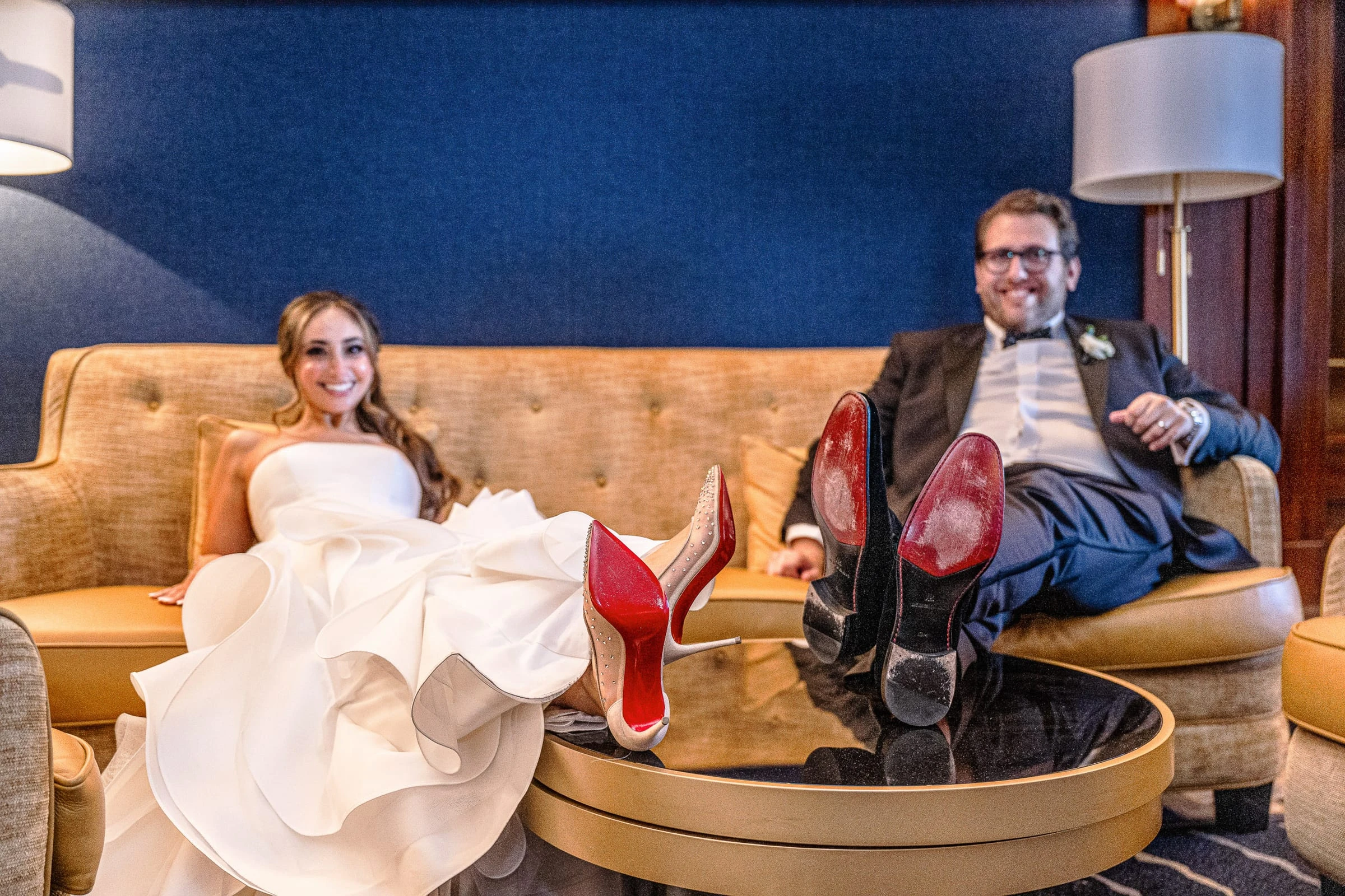 A bride and groom laughing on a sofa with their red-soled shoes propped on the table