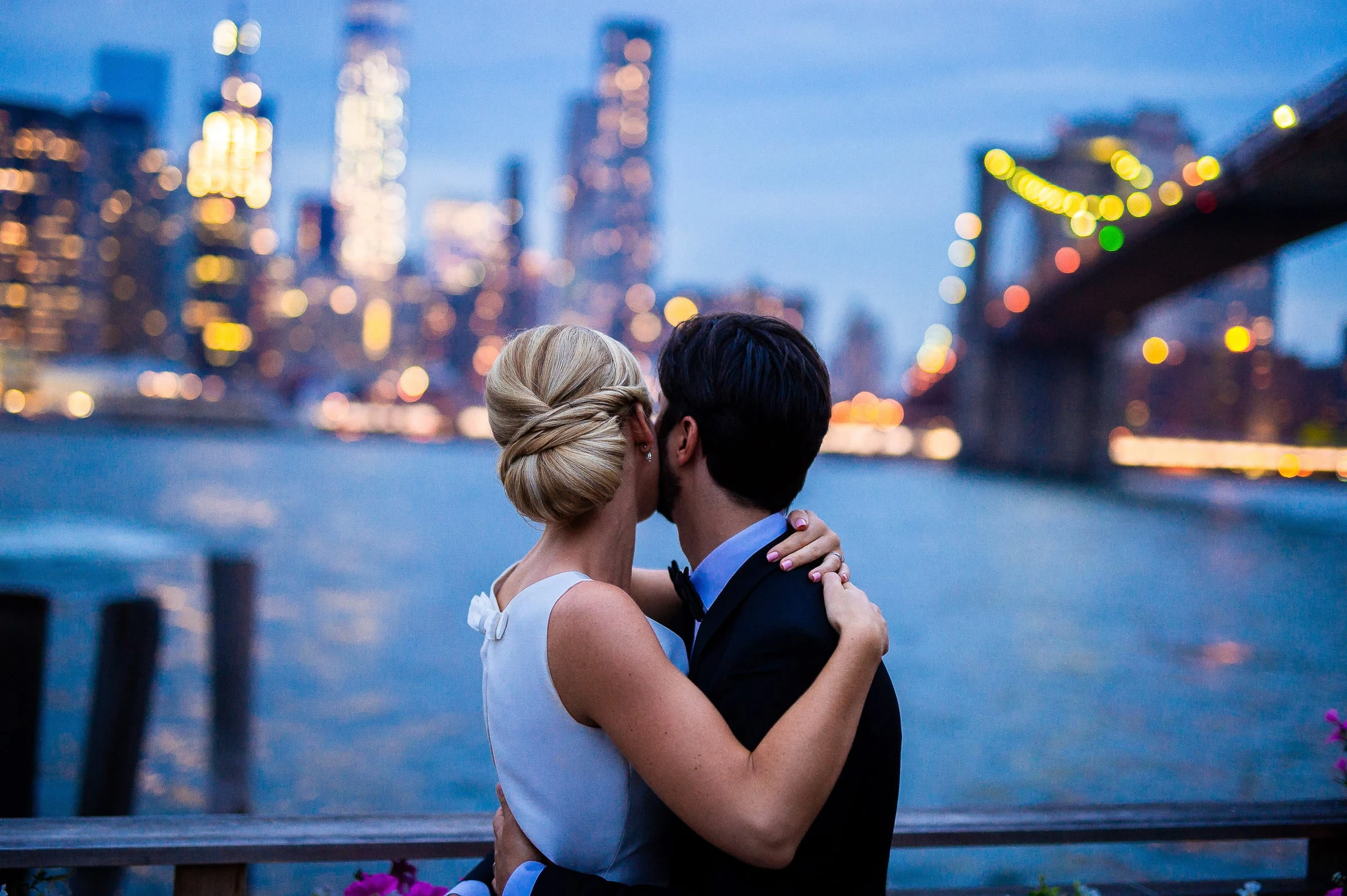 A couple kissing at a railing at dusk against the lit Manhattan skyline and the Brooklyn Bridge