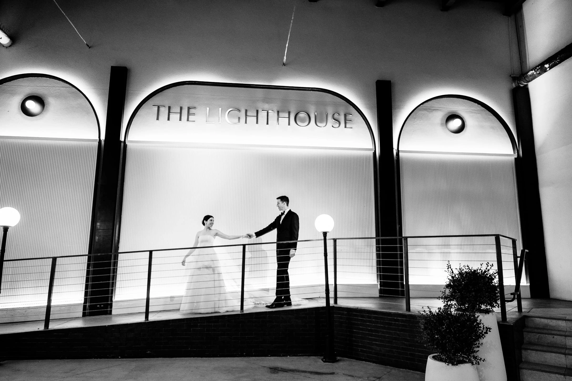 A couple meeting hands on a terrace beneath a lit sign reading The Lighthouse, in black and white
