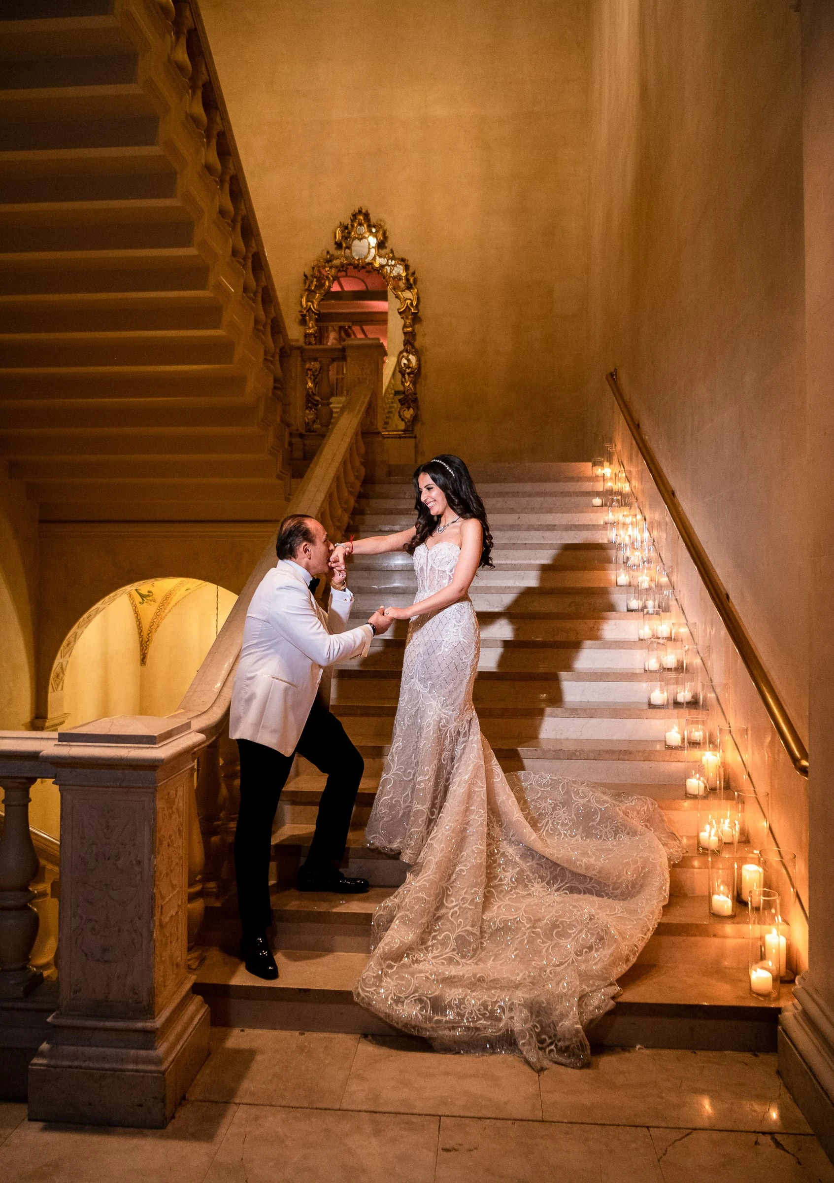 A groom kneeling to adjust the bride's train on a candlelit staircase