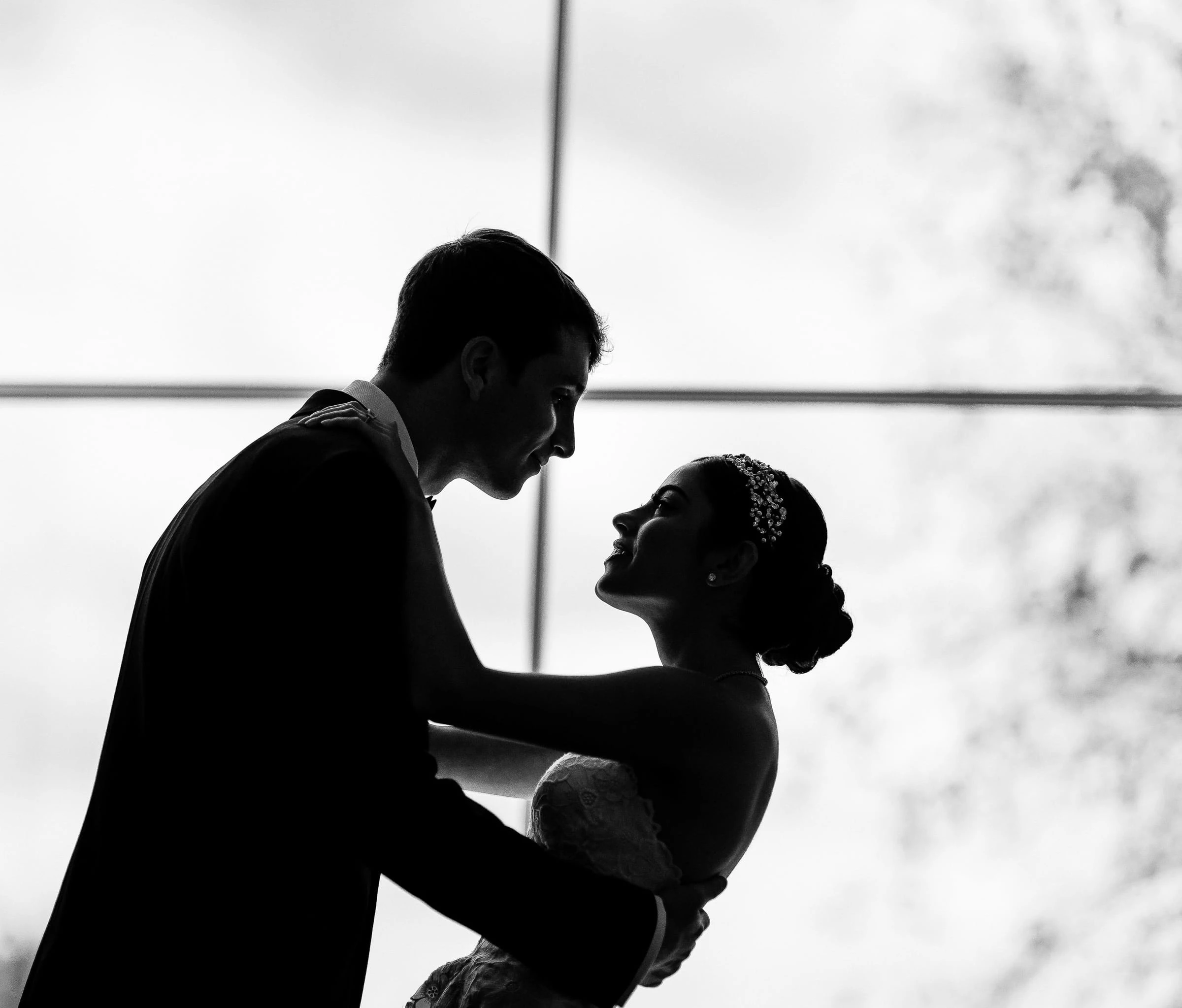 A groom leaning toward his bride in silhouette against a bright window, in black and white