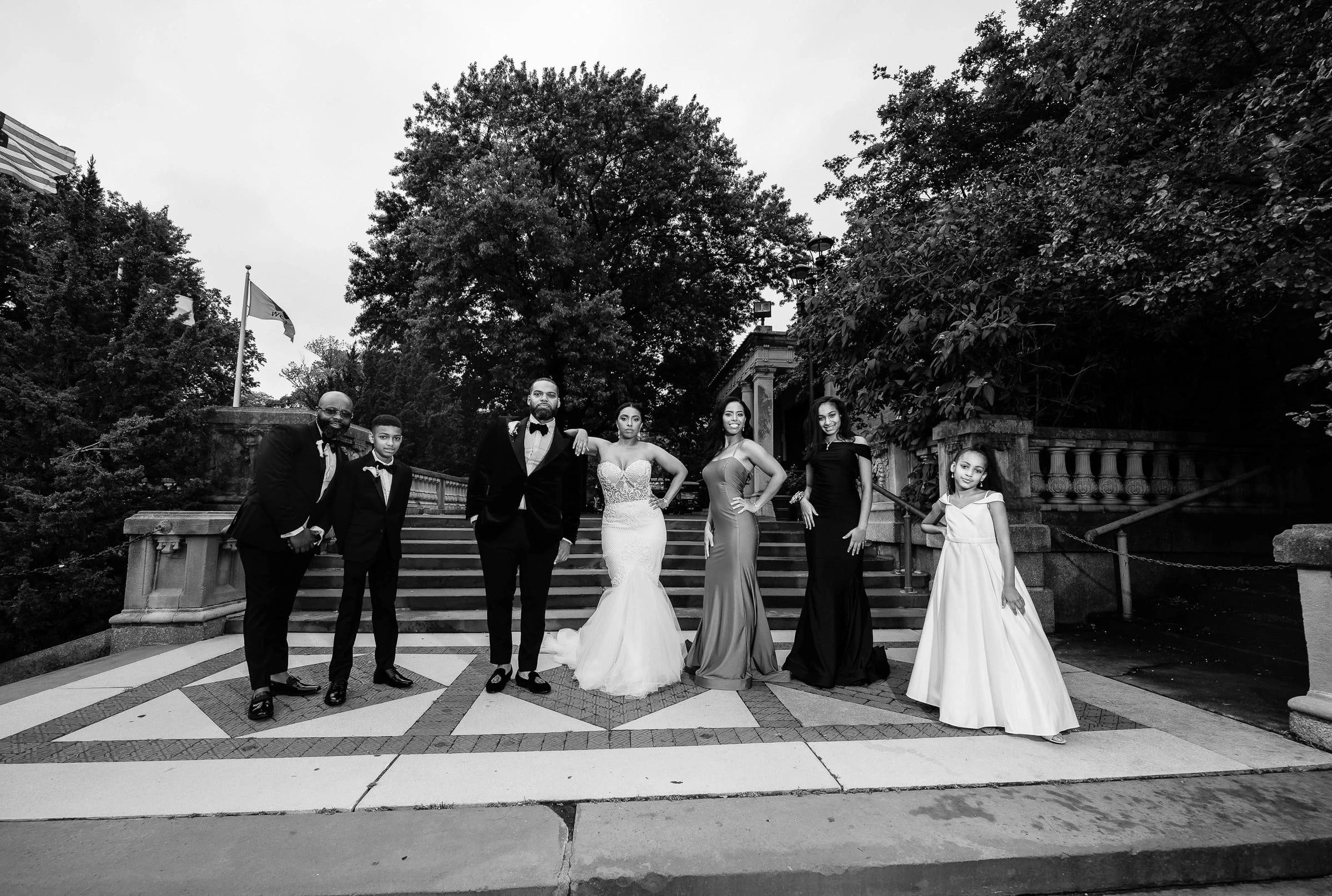 A wedding party posed on stone terrace steps beneath tall trees, in black and white