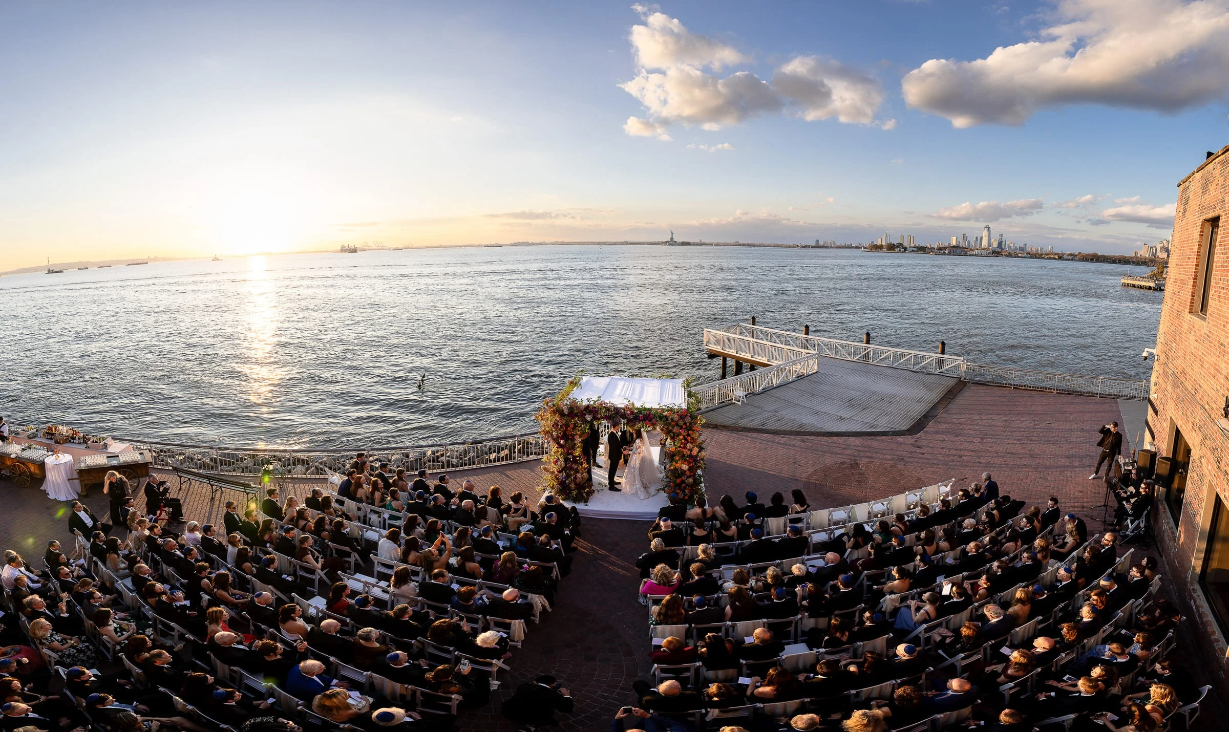 A waterfront ceremony at sunset seen from above, guests facing the chuppah by the water
