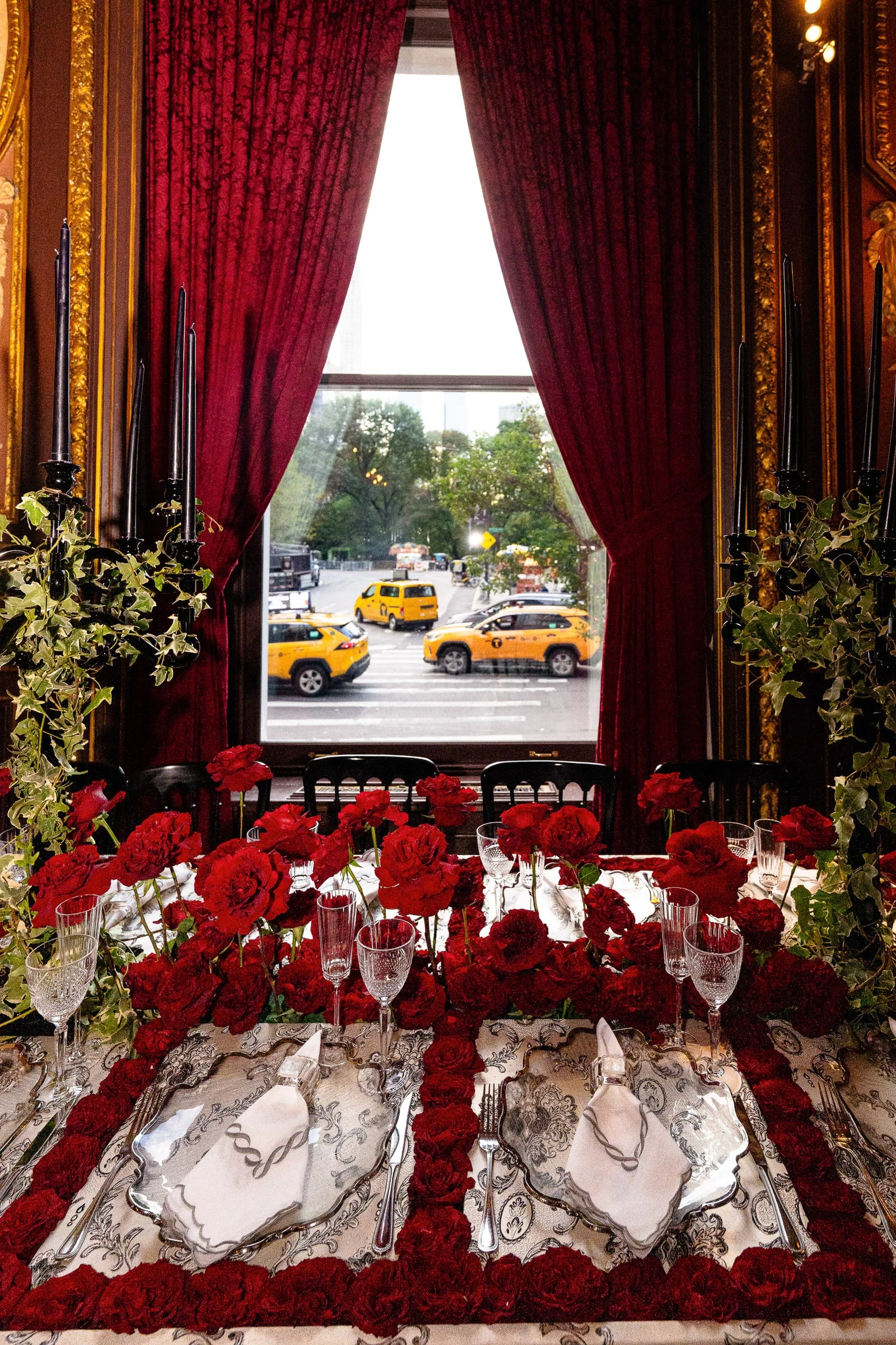 A table set with deep red roses beside a curtained window looking out onto yellow cabs