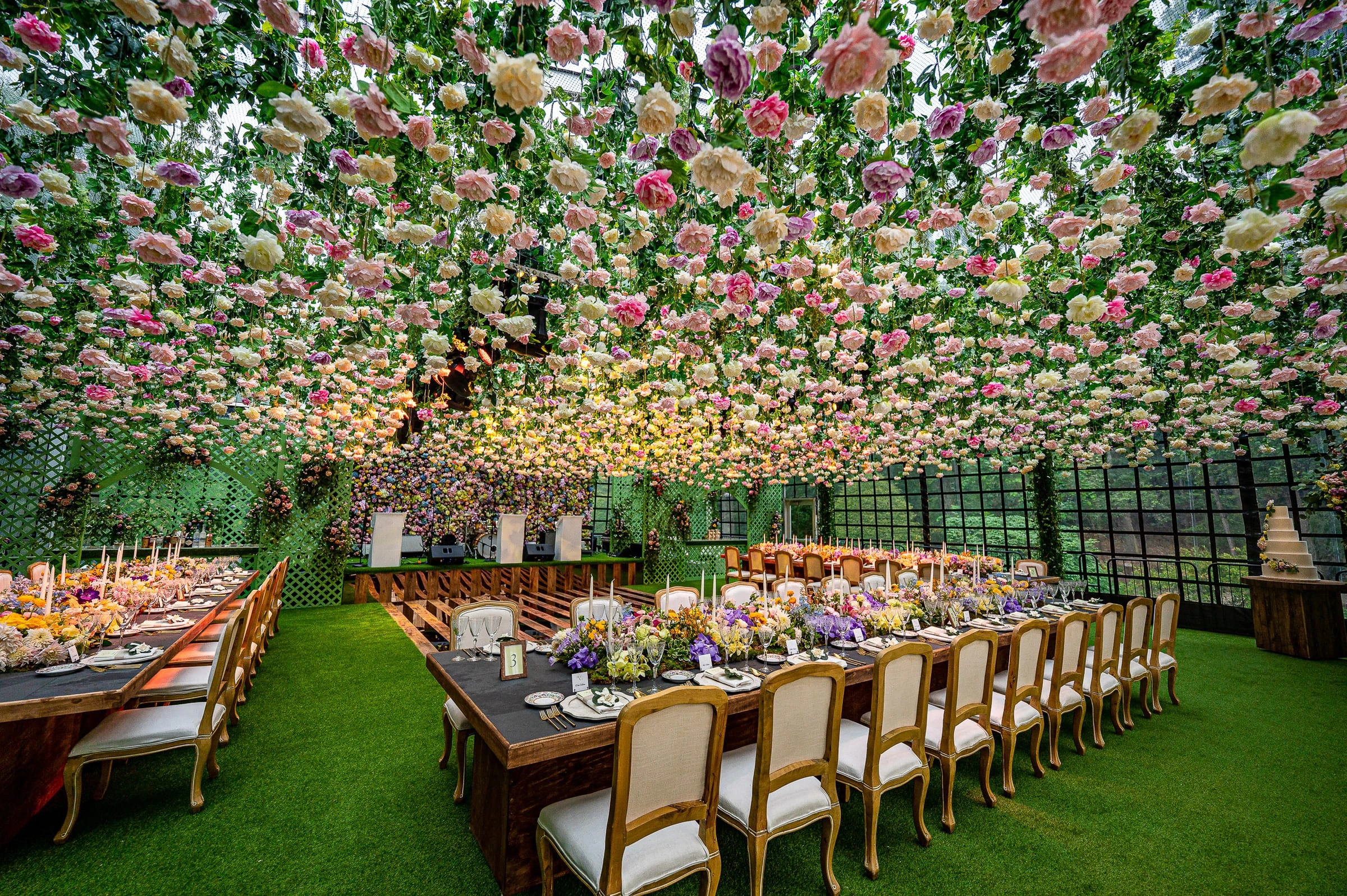 Long banquet tables on lawn turf beneath a ceiling packed with pastel roses in a trellised garden room