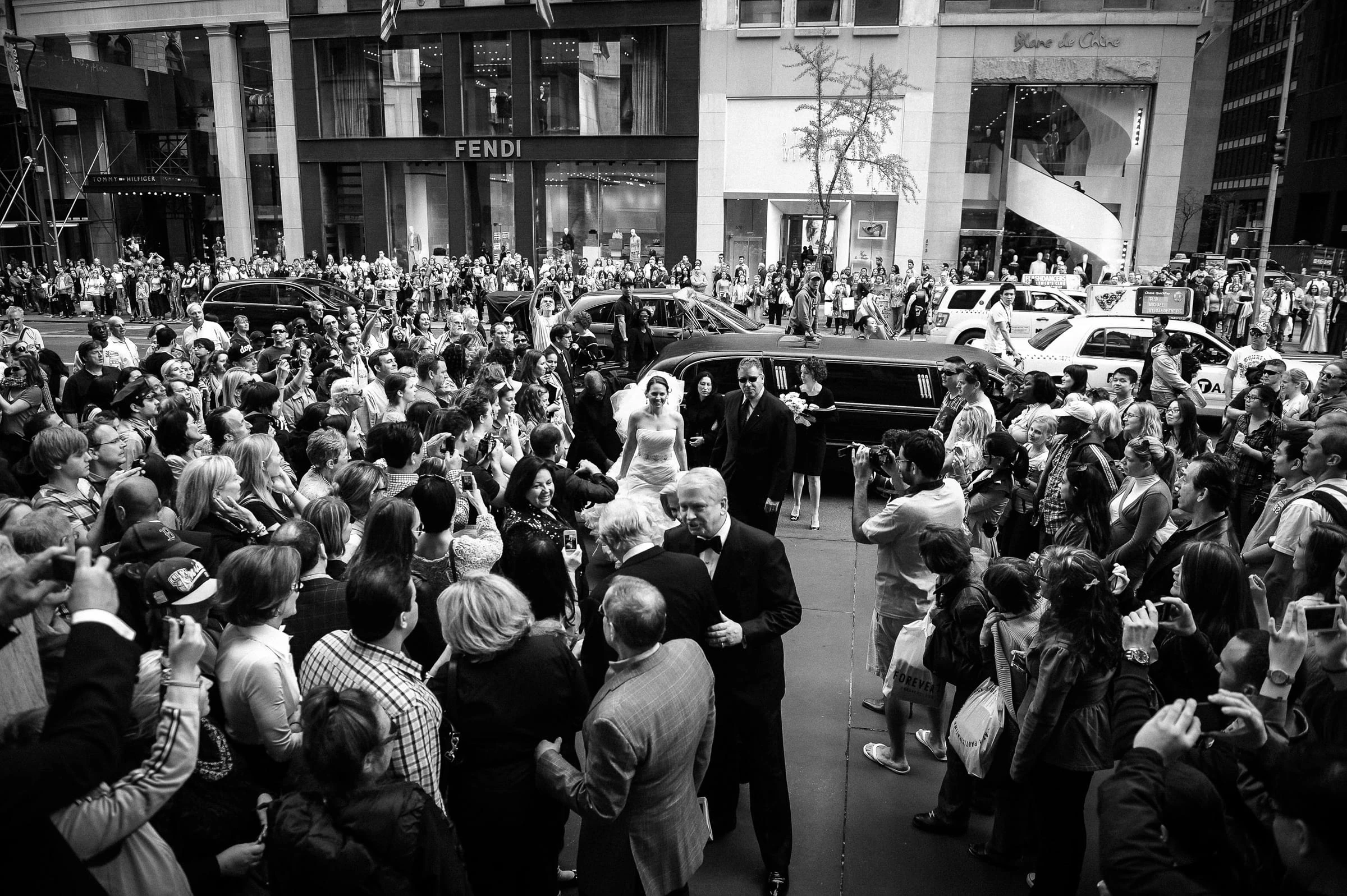 A couple arriving through a dense crowd on a city avenue, in black and white