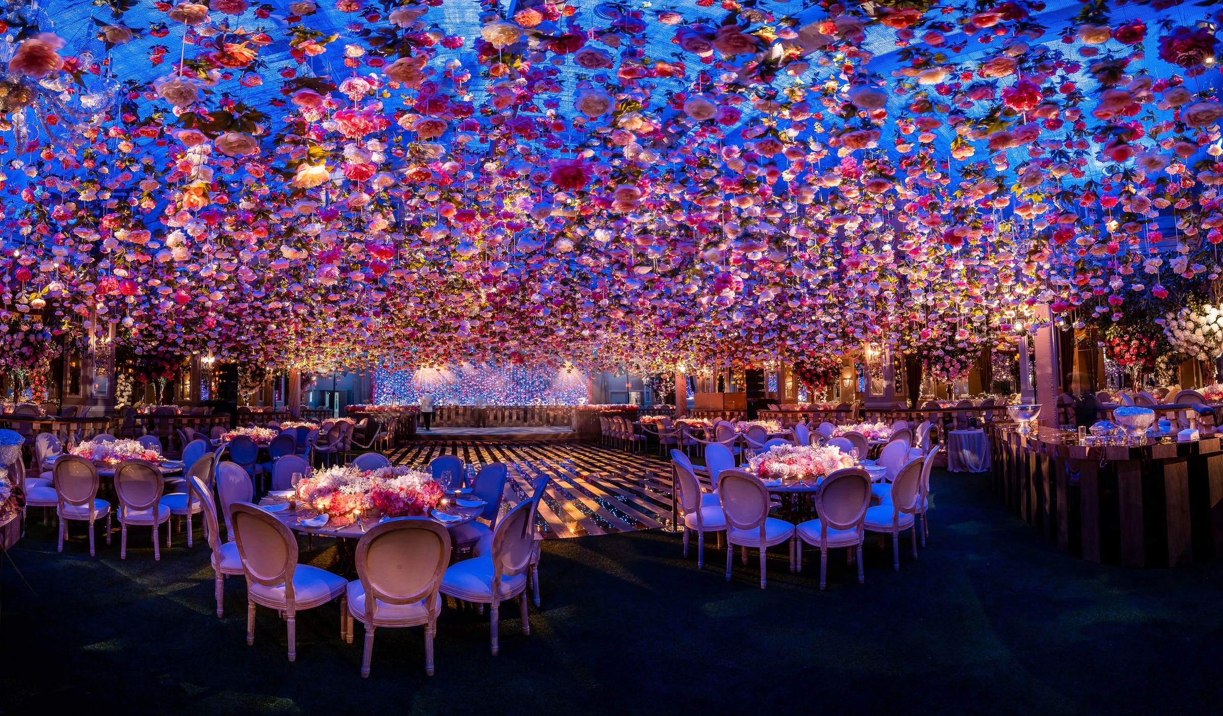 A ballroom of round tables beneath a vast ceiling of suspended flowers lit in blue and pink