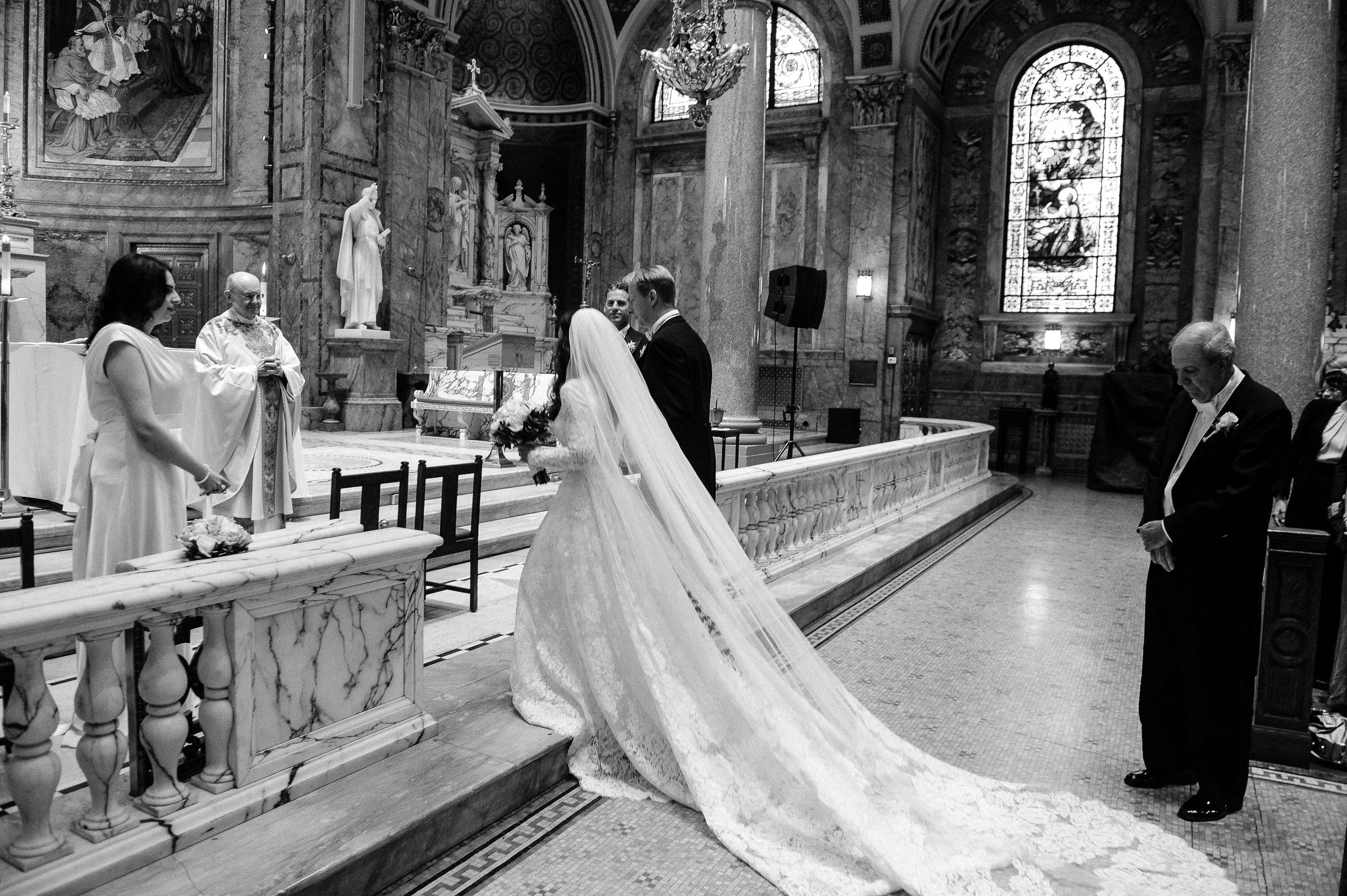 A bride with a long train at the altar rail of a church, in black and white