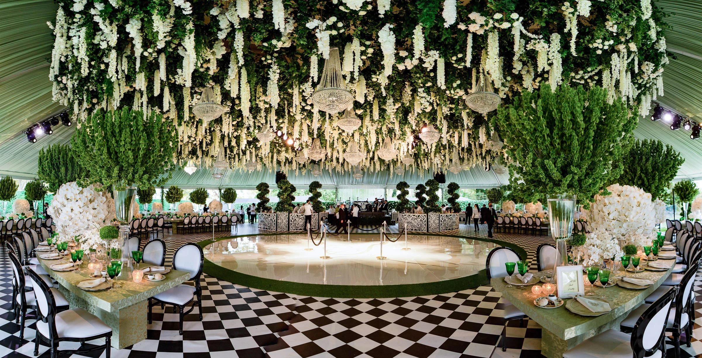 A checkerboard dance floor ringed by topiary beneath hanging greenery and chandeliers in a tented room