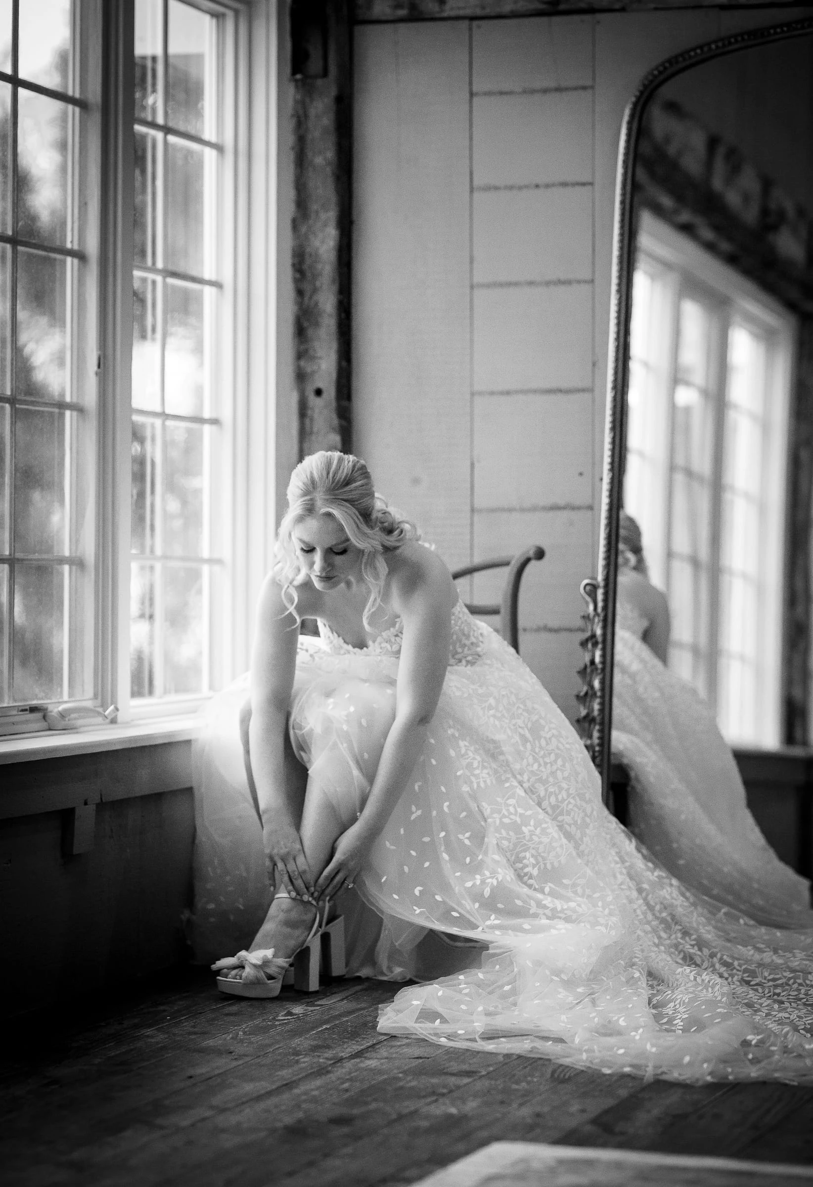 A bride fastening her shoe by a tall window, in black and white