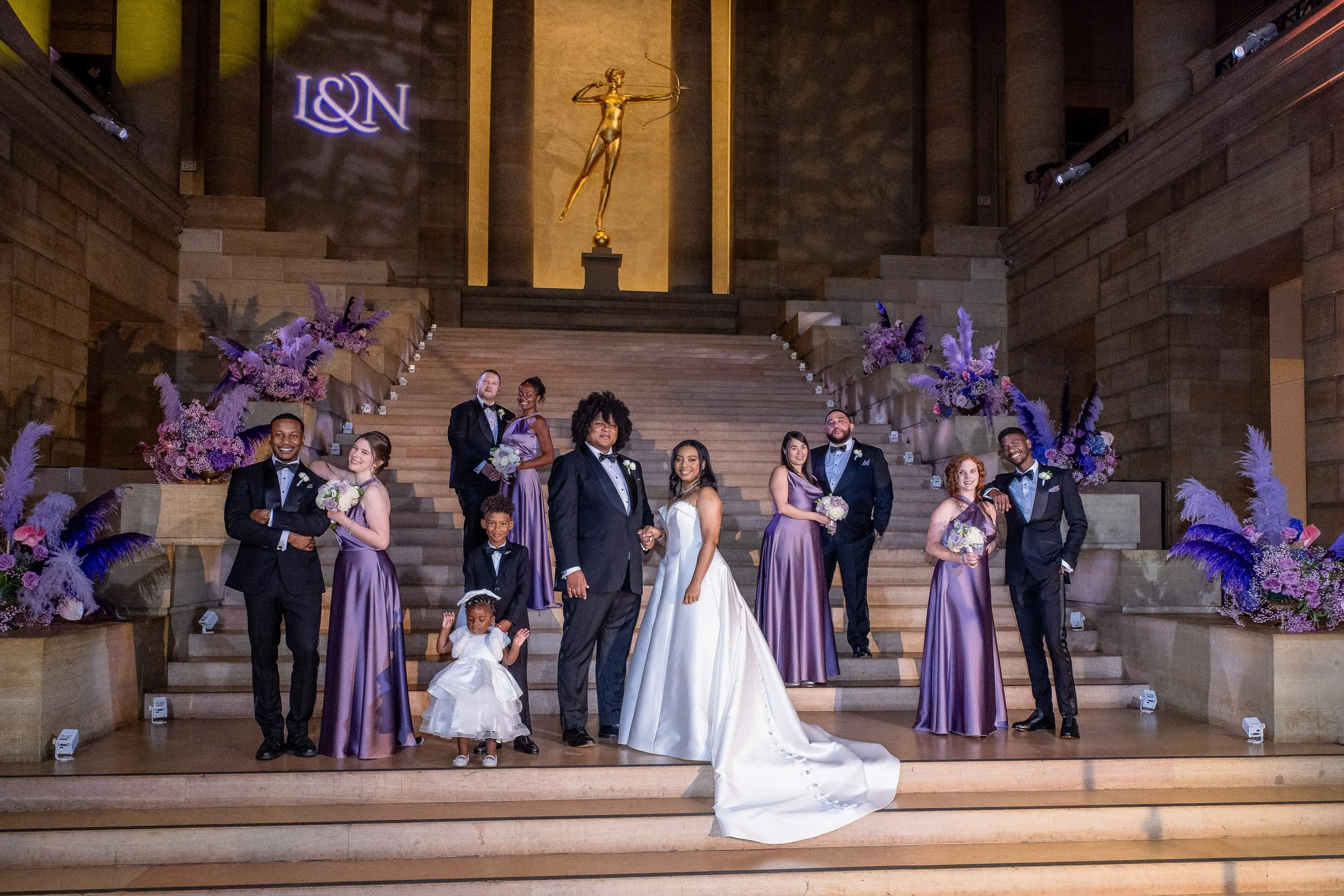 A wedding party posed on a grand museum staircase under purple lights