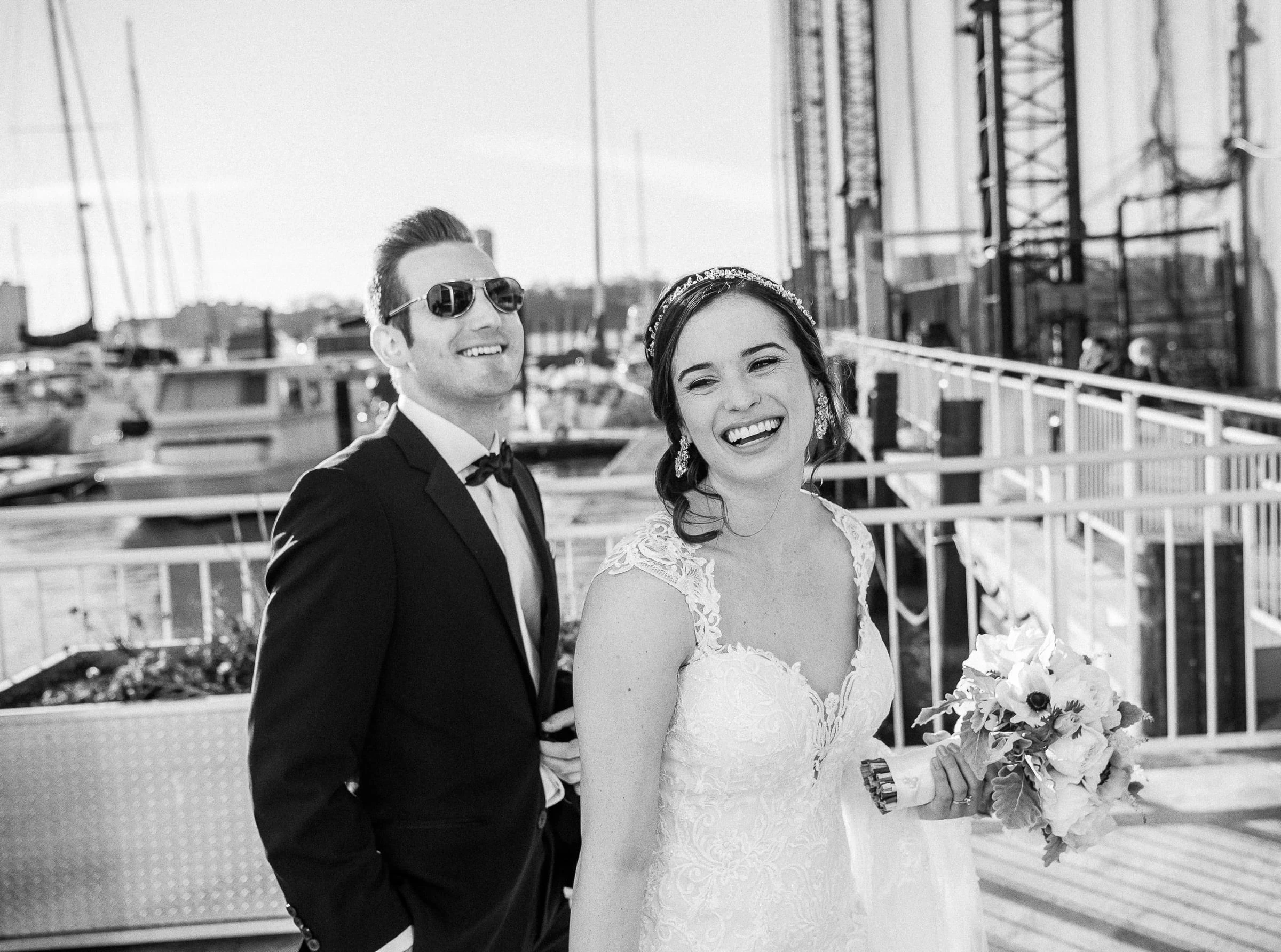 A groom in sunglasses and his laughing bride on a terrace above a marina, in black and white