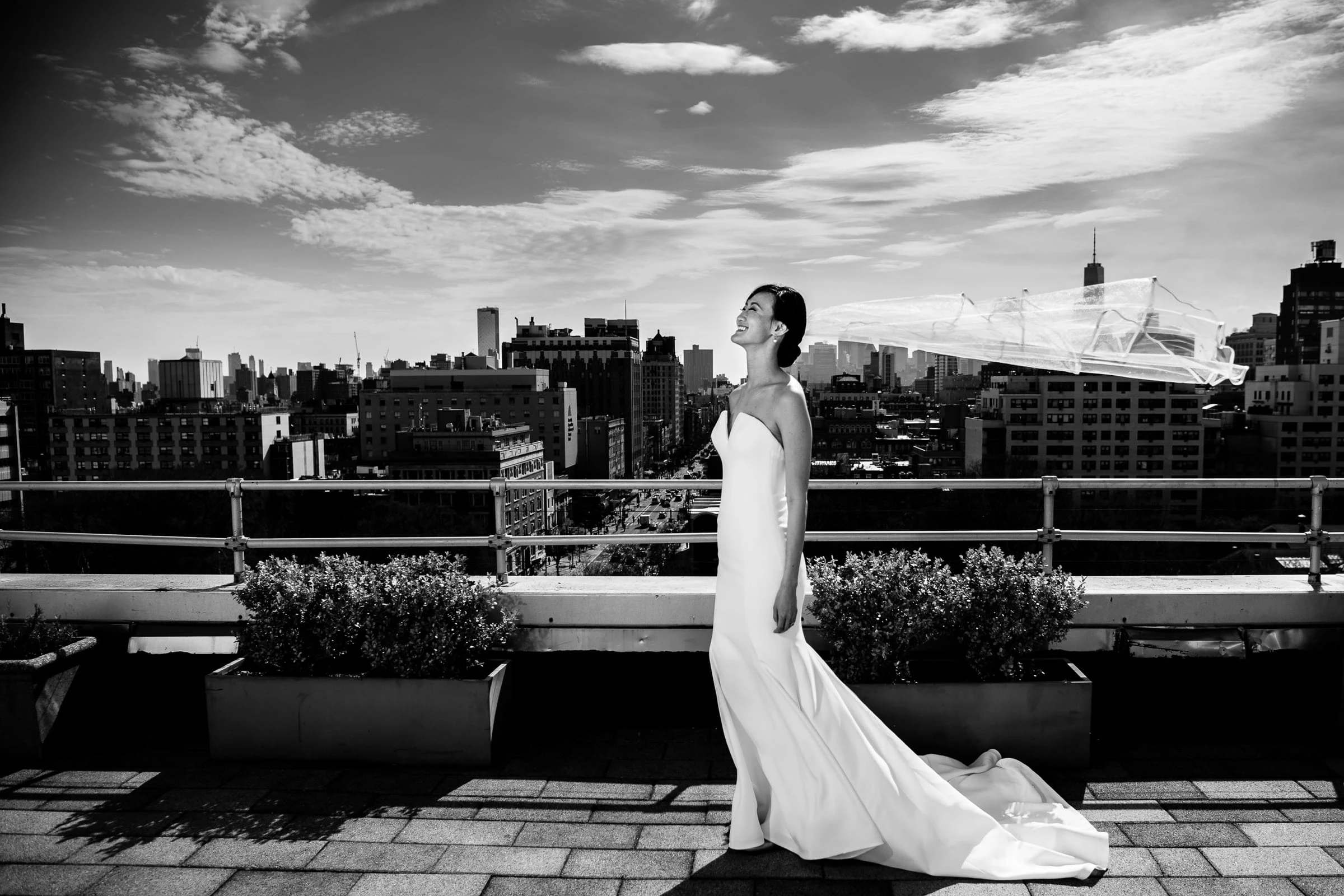 A bride on a rooftop terrace with her train spread behind her and the city skyline beyond, in black and white