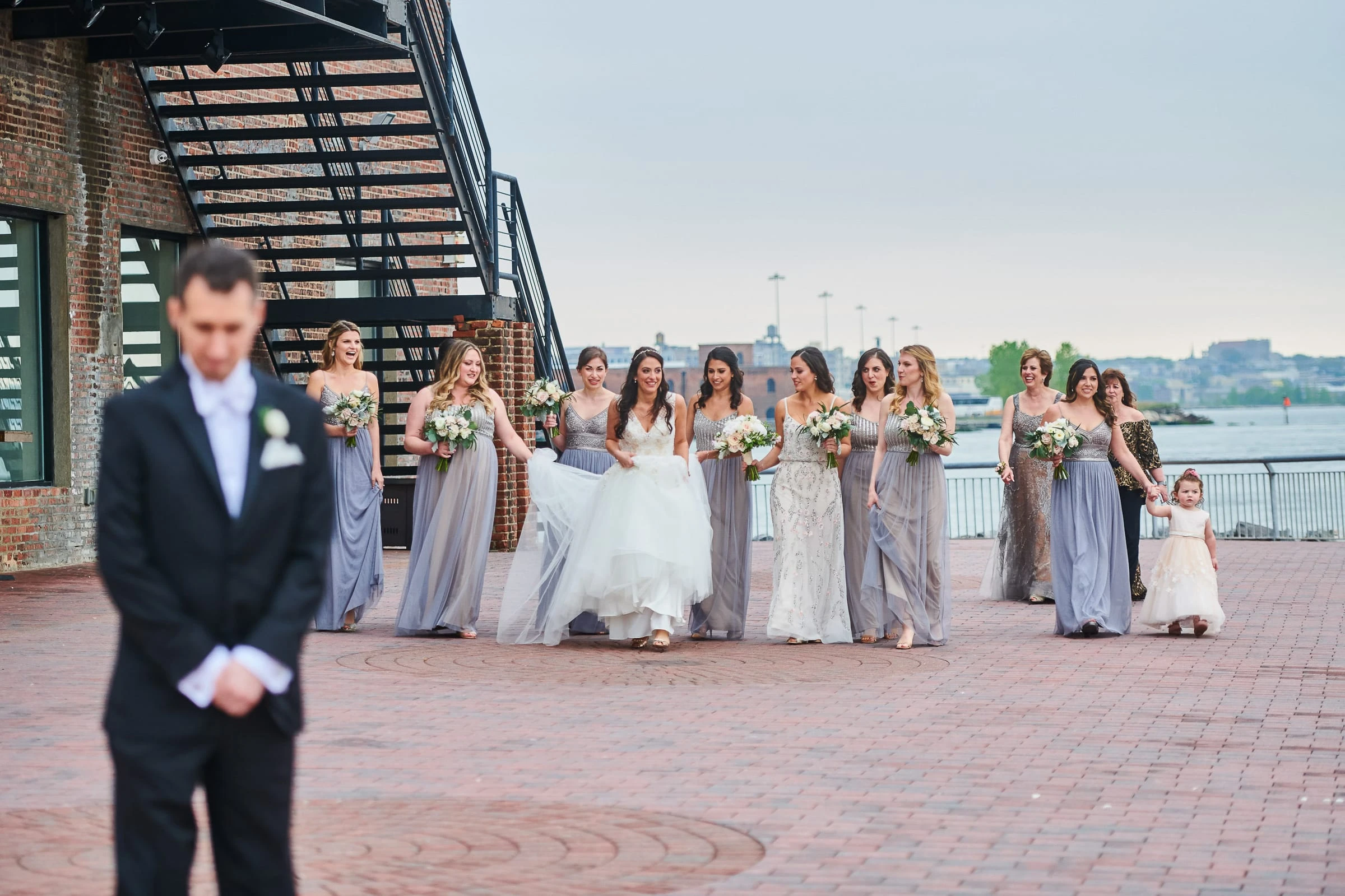 A groom waiting with his back turned as the bride approaches with her bridesmaids on a waterfront terrace