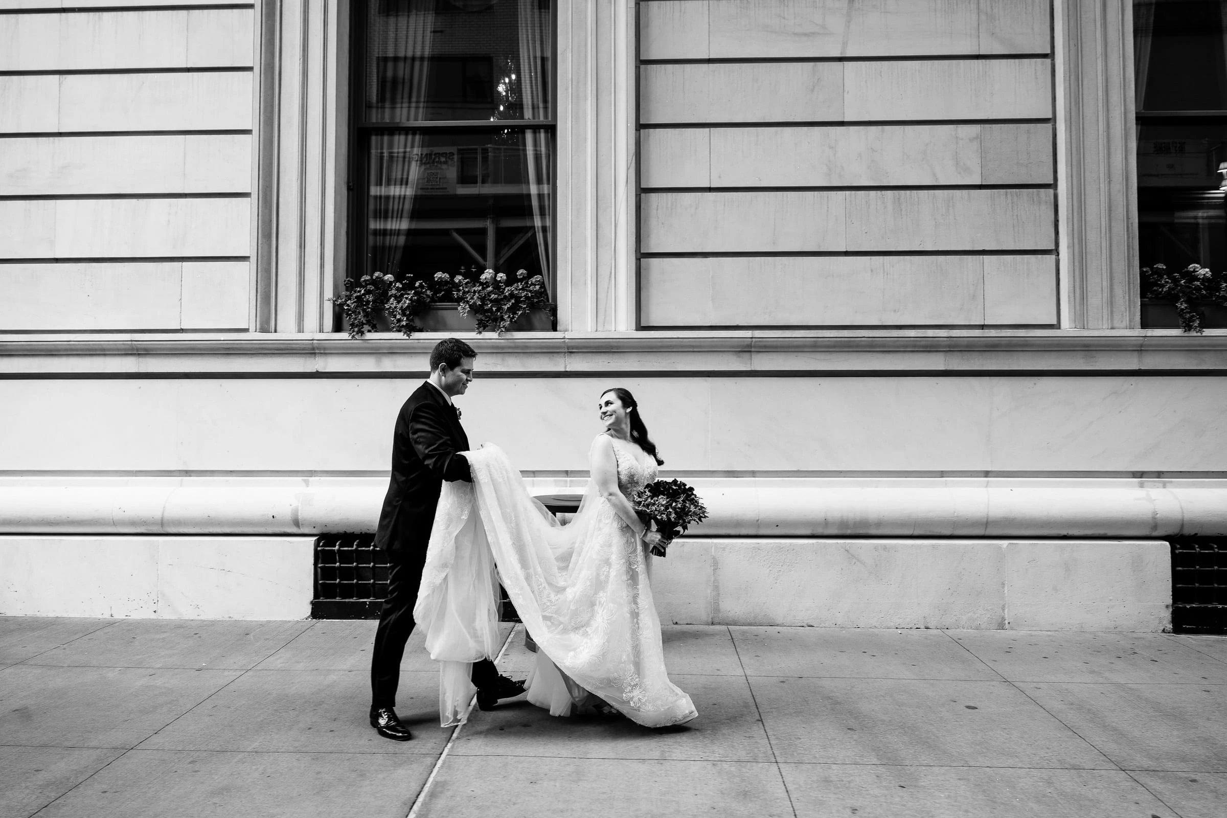 A couple walking past a stone building, the groom carrying her train, in black and white