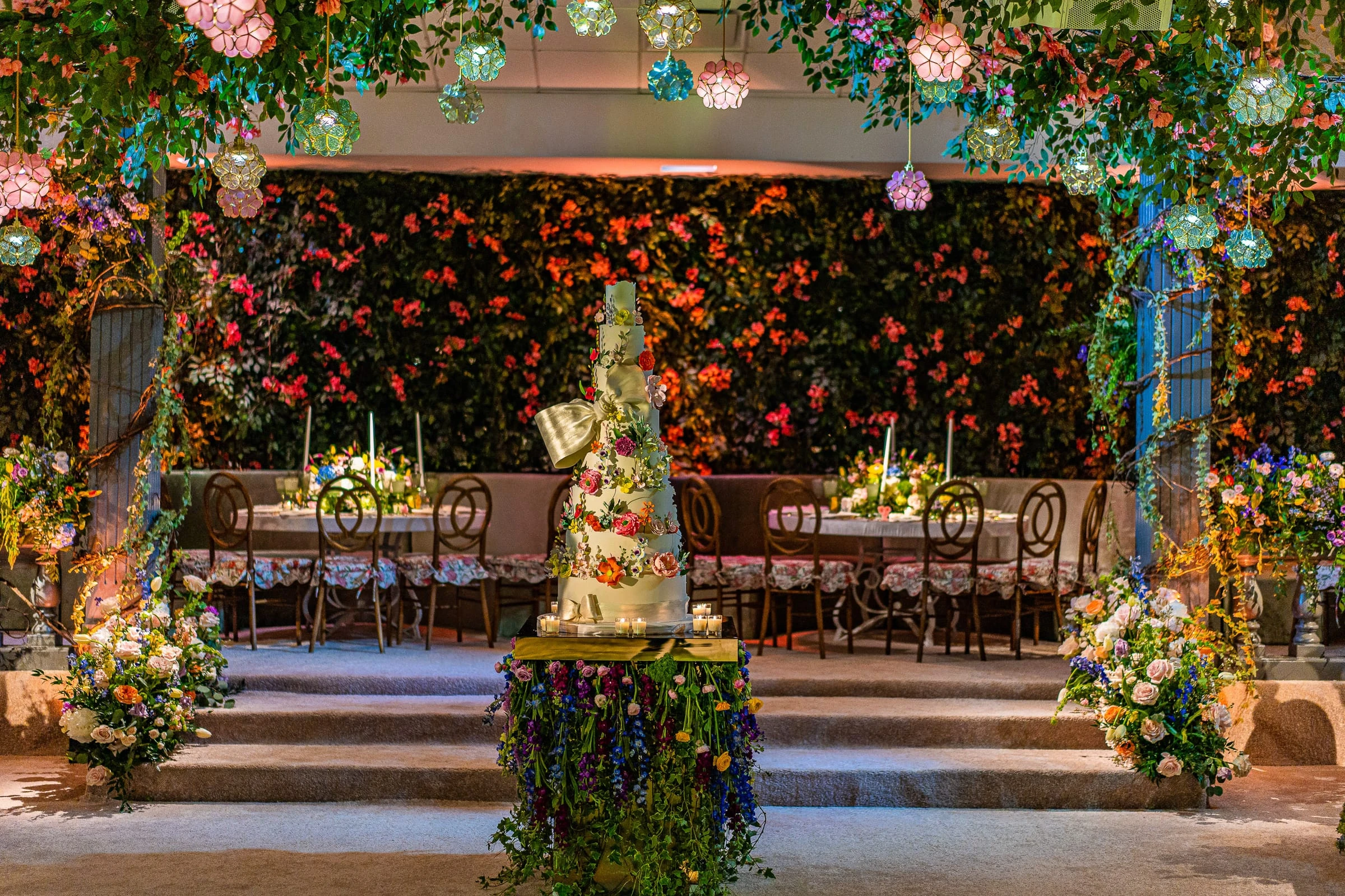 A tiered cake in front of a wall of wildflowers and long tables in a garden-styled room