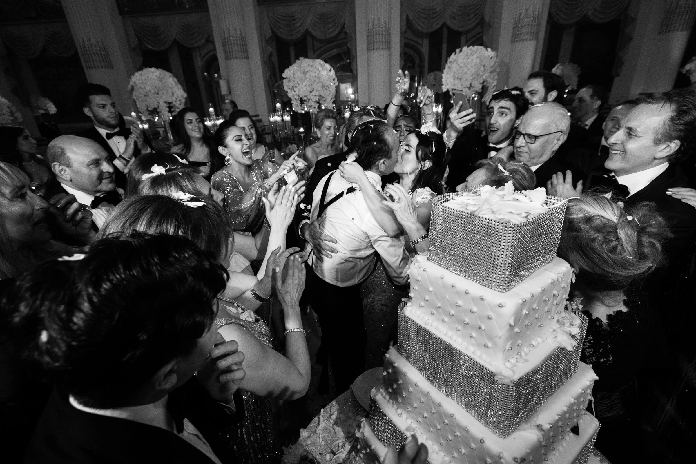 Guests crowding around a tall wedding cake in celebration, in black and white