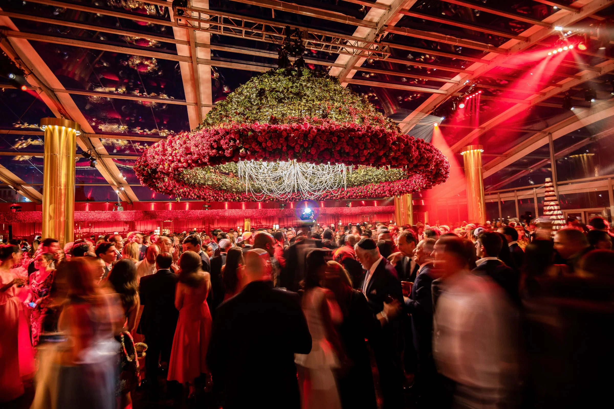 A packed reception in red light beneath a hanging ring of flowers