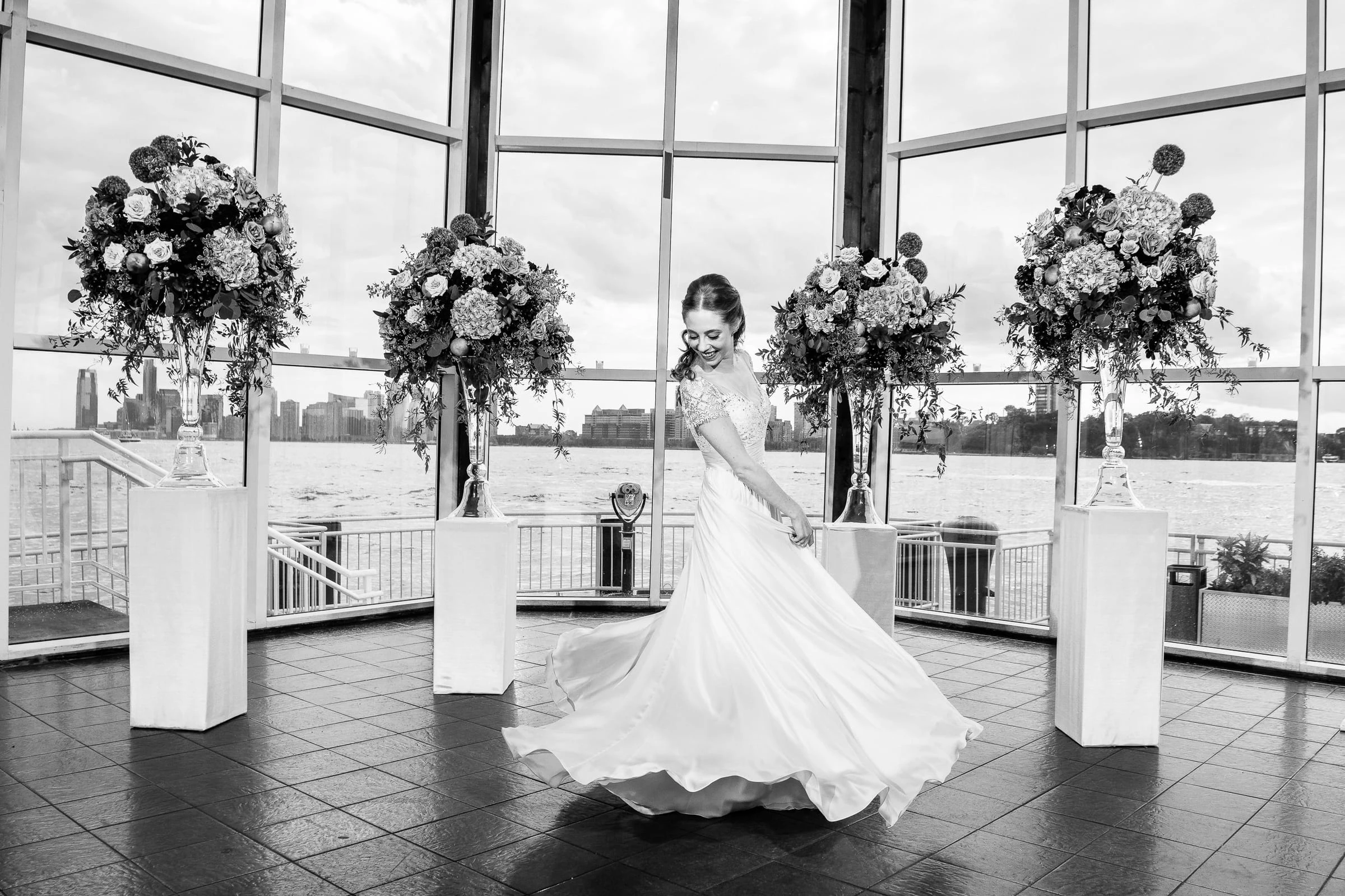 A bride twirling her skirt between tall flower arrangements by the windows, in black and white