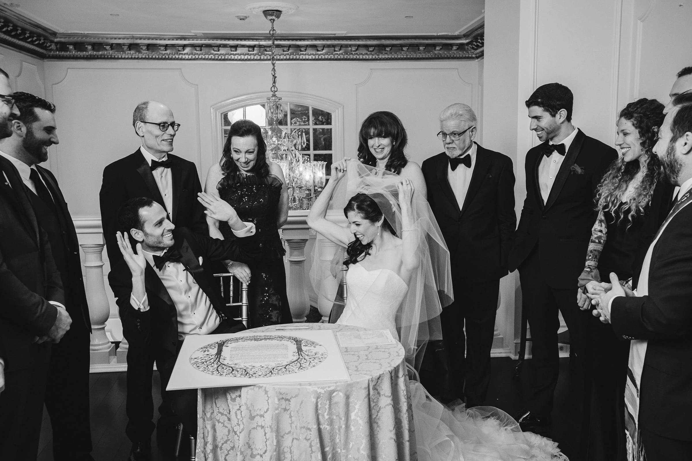 A groom signing the ketubah as the bride and family watch, in black and white