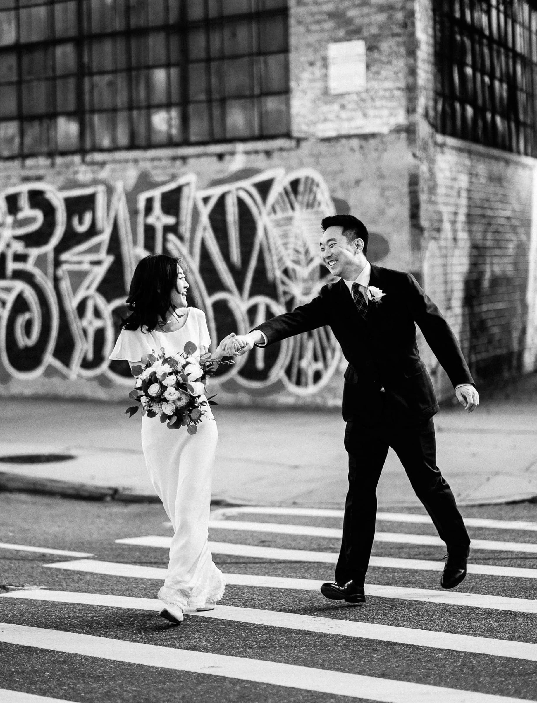 A couple crossing the street hand in hand past a graffiti wall, in black and white