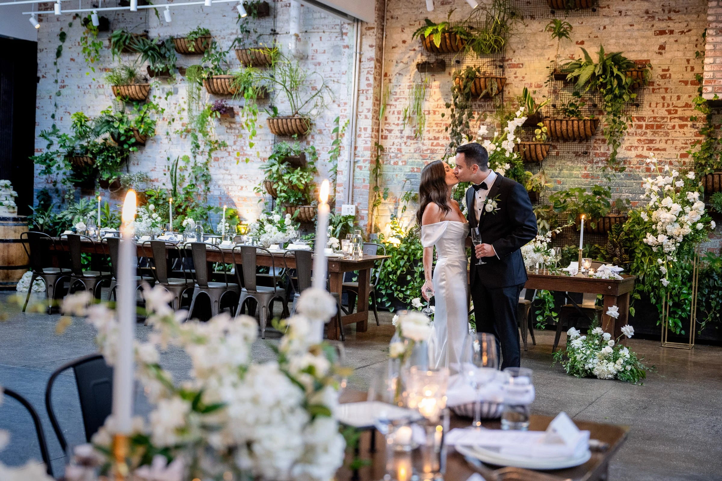 A couple embracing among candlelit tables beneath a wall of hanging plants