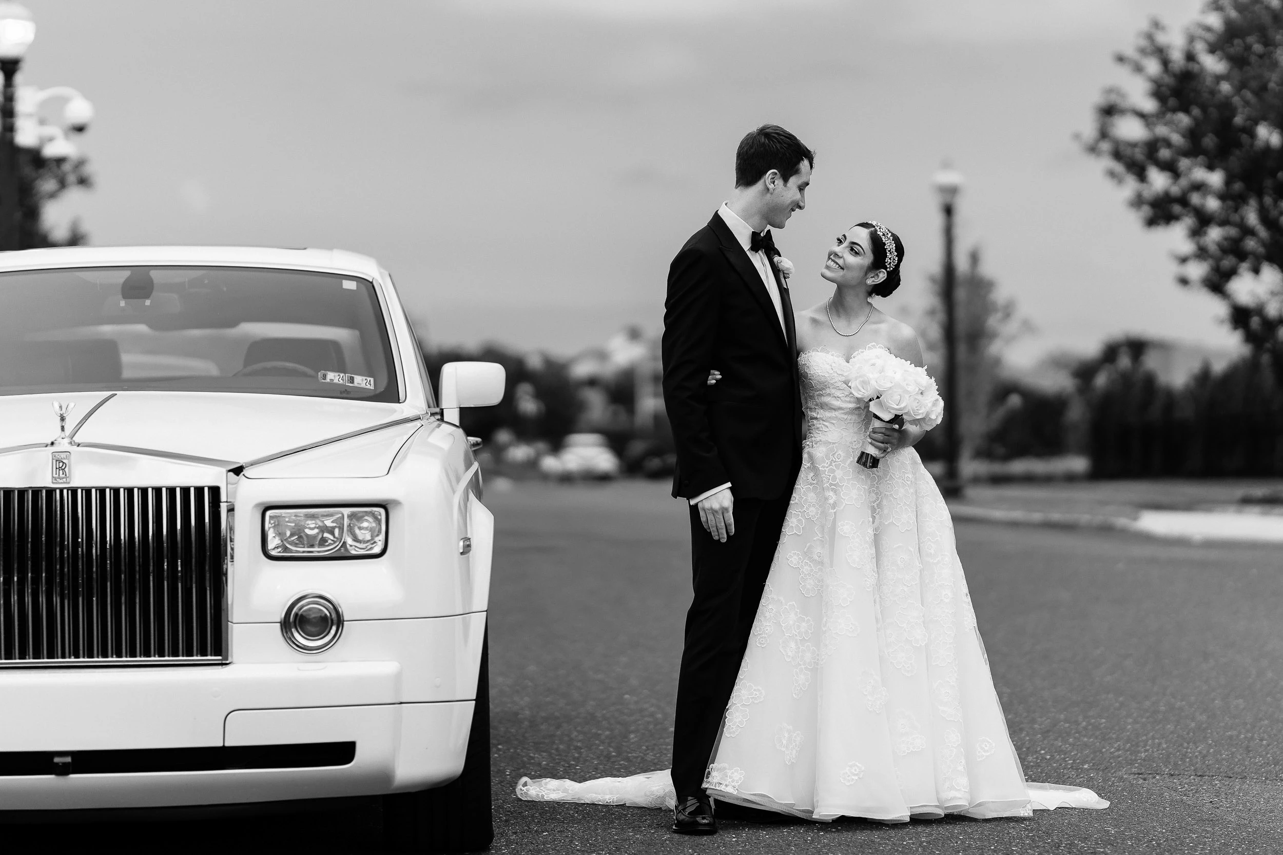 Black and white photograph of a bride and groom beside a white Rolls-Royce Phantom on a lamplit drive.